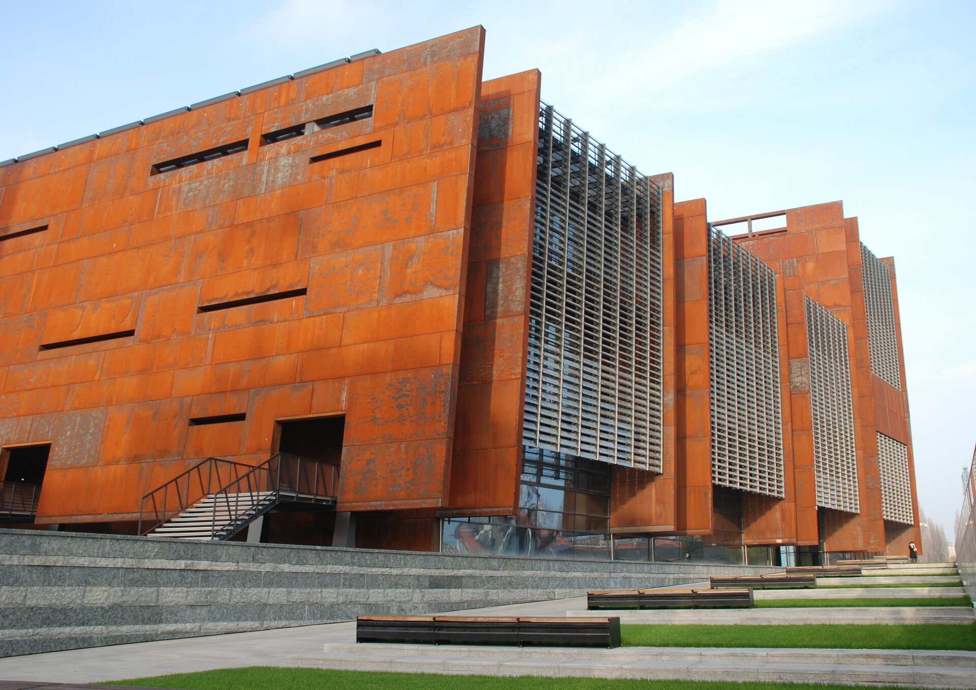 Fotografia przedstawiająca Europejskie Centrum Solidarności w Gdańsku. Budynek z czerwonej cegły z aluminiowymi akcentami. Kubatura budynku nowoczesna. Budynek otoczony betonowym chodnikiem poprzecinanym trawnikami.