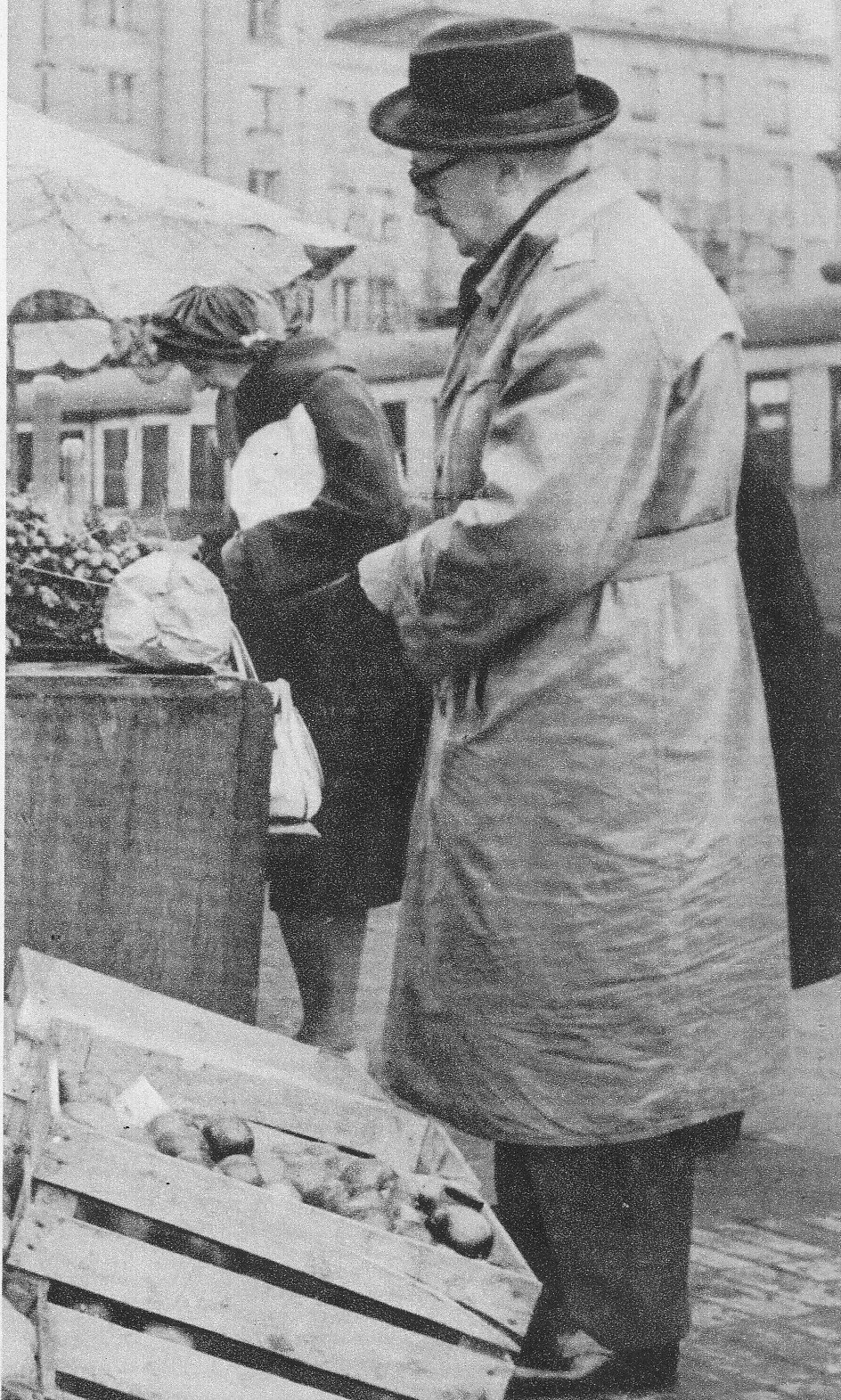 Fotografia przedstawia dojrzałego mężczyznę stojącego na ulicy, obok straganu. Postać prezentuje lewy profil. Mężczyzna ma duży nos i małe wąsy. Nosi okulary w grubej oprawie. Na głowie ma kapelusz, Jest ubrany w płaszcz. Pod stopami mężczyzny jest ustawiona drewniana skrzynka z jabłkami. Za nimi stoi prowizoryczna lada, na której leży główka kapusty. Na drugim planie ukazana jest kobieta pochylająca się nad warzywami. W tle tramwaj i kamienica.