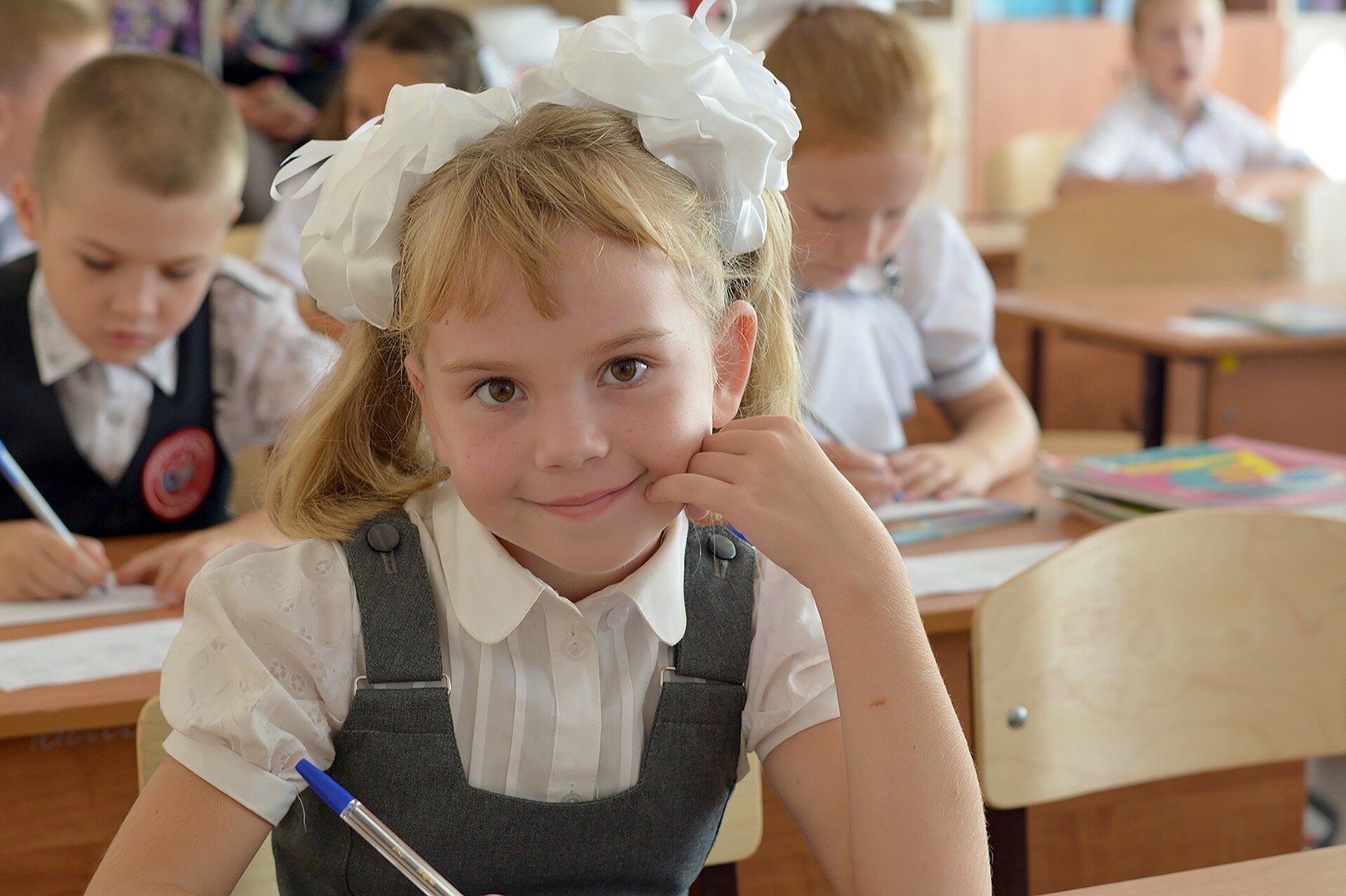 Zdjęcie przedstawia uśmiechniętą dziewczynkę. Dziewczynka siedzi przy szkolnej ławce i trzyma w ręku długopis. Za nią przy ławkach siedzą inne dzieci, które piszą na kartkach.