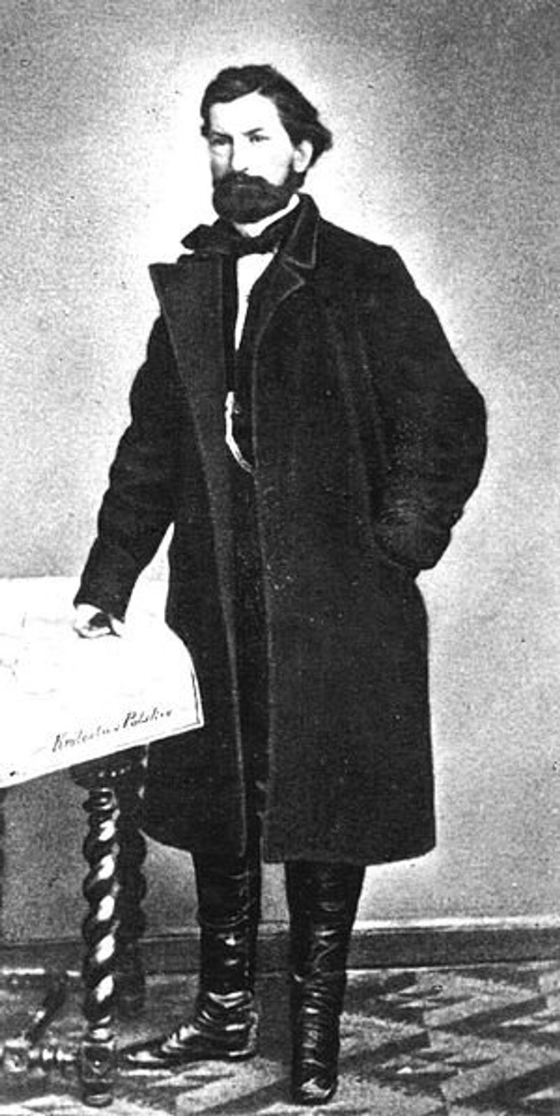 Kliknij, aby powiększyć Zdjęcie przedstawia postać Marcina Borelowskiego „Lelewela”, stojącego przy stoliku nakrytym obrusem i posiadającym rzeźbione nogi. Jego prawa dłoń jest oparta o stół, a lewa znajduje się w kieszeni wełnianego płaszcza. Mężczyzna ubrany jest w wysokie buty, koszulę z wysokim kołnierzem i chustą pod szyją. Ma zaczesane do tyłu włosy oraz wąsy i brodę.