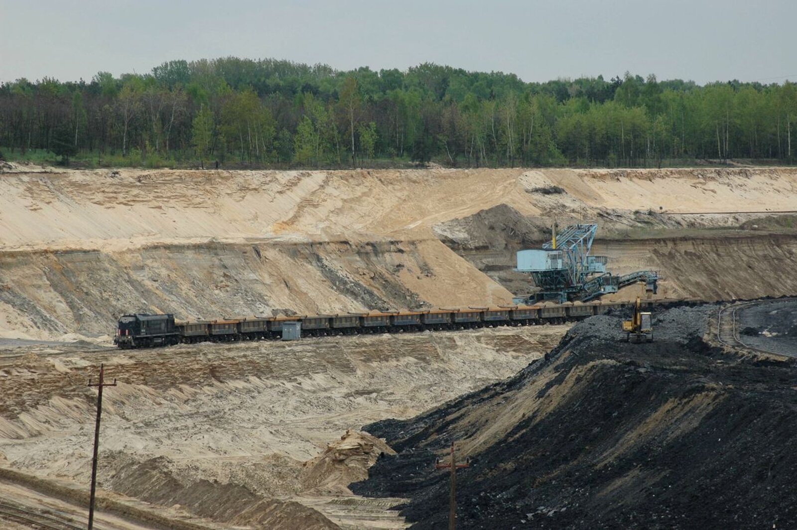 Fotografia kopalni piasku. Jest dużych rozmiarów, tworzy zagłębienie o wielometrowej głębokości. Pośrodku podstawiony pociąg z wagonami, na które wsypywany jest piasek za pomocą ogromnej koparki stojącej obok.