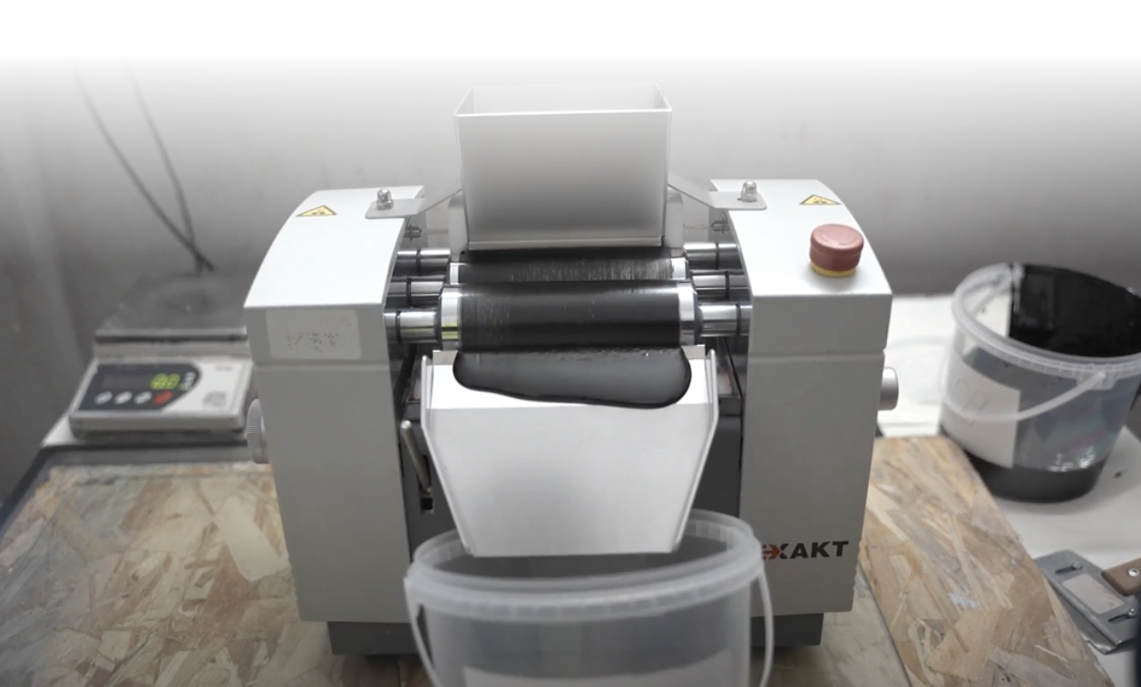 Zdjęcie przedstawia walcarkę. To nieduże urządzenie z systemem trzech wąskich walców, które formują masę w równy plaster. Przed walcarką znajduje się nieduże wiadro, do którego zsuwa się po pochyłej tacy walcowana masa.