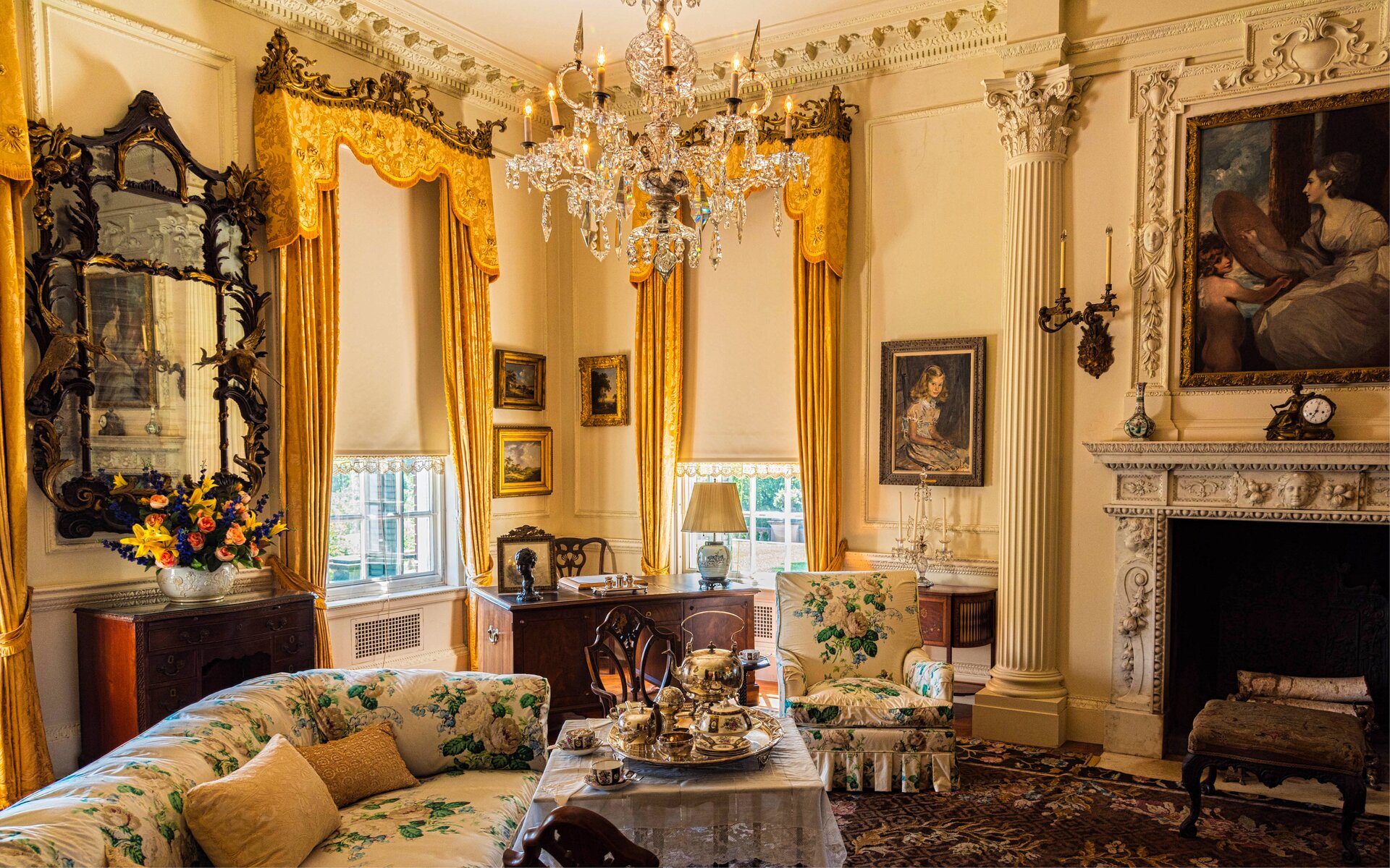 Obrazek pokazuje salon w stylu historycznym. Jasne, eleganckie wnętrze wypełnia ciepłe światło wpadające przez wysokie okna pięknie zdobione zasłonami. Ściany w beżowych kolorach są udekorowane kolumnami, a blisko sufitu znajduje się sztukateria. Główną część zajmuje bogato zdobiony kominek, na którym stoi elegancki zegar. Na jasnej podłodze leży wzorzysty dywan, a centralnym punktem jest sofa w jasnym materiale z delikatnymi kwiatowymi wzorami i stojący obok niski stolik z porcelaną. Nad szafką pomiędzy oknami wisi duże lustro w ozdobnej ramie, a na ścianach zawieszono, olejne obrazy w eleganckich ramach. W rogu salonu pod oknem stoi biurko z krzesłem. Na suficie wisi żyrandol z kryształami, tworząc elegancki akcent w całym salonie.