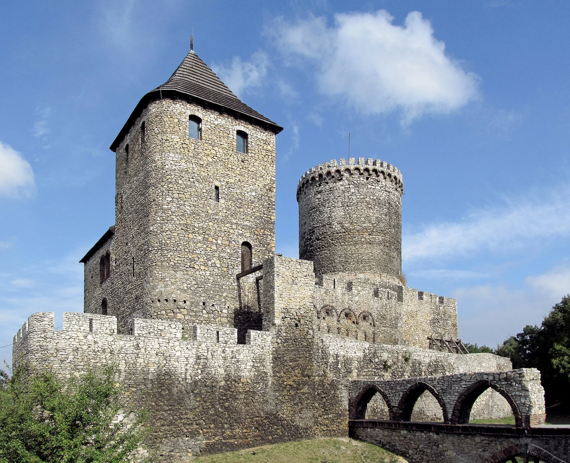 Zdjęcie przedstawia zamek w Będzinie. Zamek oraz mury obronne zostały wykonane z licznych kamieni, tworzących gruby mur. Do wysokiej, kwadratowej wieży ze spiczastym, drewnianym dachem, dobudowano niższą część. W wieżę wbudowano małe, półkoliste okna oraz drewniane drzwi. Po prawej stronie znajduje się okrągła wieża obronna, nieco niższa od drugiej wieży. Całość jest otoczona grubymi murami obronnymi. Po prawej stronie fotografii znajduje się kamienny most. Wokół budowli rosną drzewa. Tło stanowi jasne niebo.