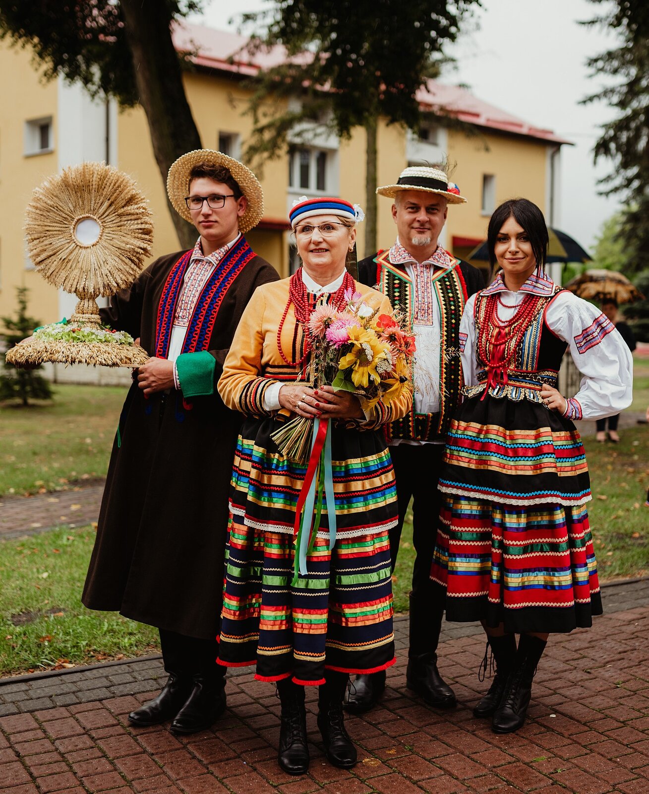 Zdjęcie prezentuje 4 osoby w ludowych strojach. Kobiety noszą pasiaste, kolorowe spódnice, gorsety oraz korale. Mężczyźni brązowe katany i plecione, słomiane kapelusze. Dwie z osób trzymają symbole. Kobieta bukiet kwiatów i zbóż, mężczyzna wyplecioną ze zboża figurę. 
