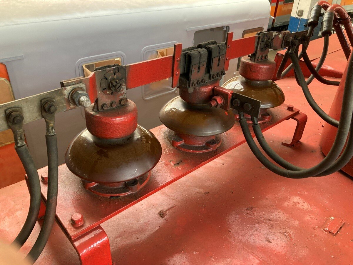 Zdjęcie przedstawia odłącznik pantografów na dachu wagonu. Na ramie umieszczona jest podstawa stalowa. Na podstawie rzędem stoją trzy izolatory, na których osadzone są styki nieruchome. Nad izolatorami znajdują się drążki ze stykami ruchomymi. Do drążków oraz powyżej środkowego izolatora podpięte są kable.