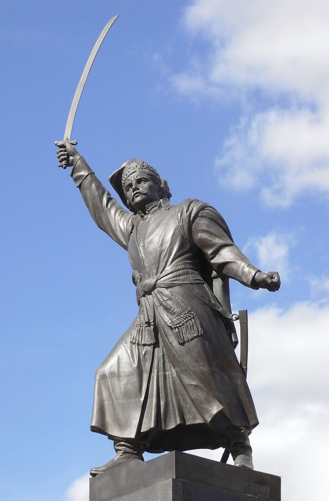 Na zdjęciu pomnik mężczyzny. W prawej dłoni ma miecz, ręka jest podniesiona do góry. Lewa ręka jest zaciśnięta. Nogi są w rozkroku. Postać ma czapkę i długi płaszcz.  