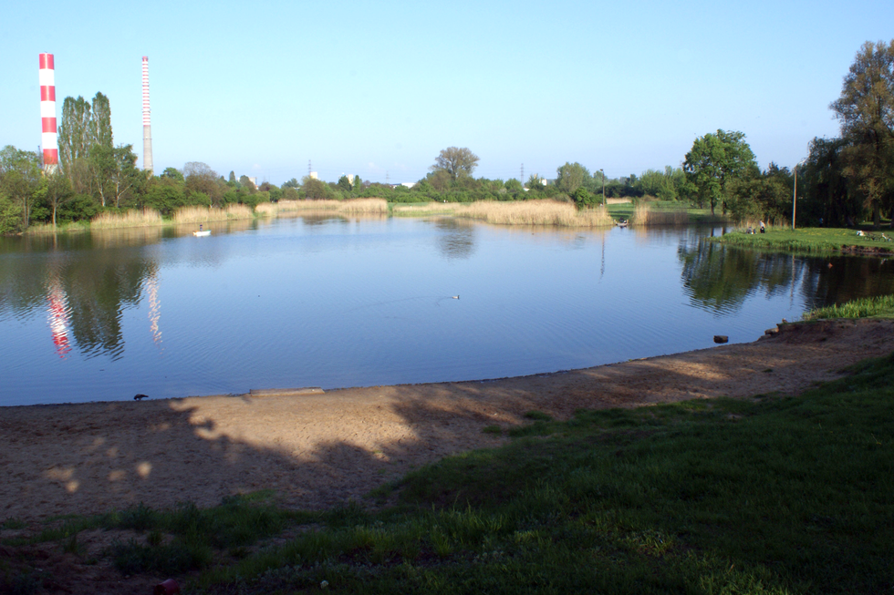 Fotografia prezentuje niewielkie Jeziorko Czerniakowskie, jezioro zakolowe powstałe ze starorzecza. Na pierwszym planie piaszczysty brzeg jeziora. Po lewej stronie za jeziorem widać kominy elektrociepłowni.