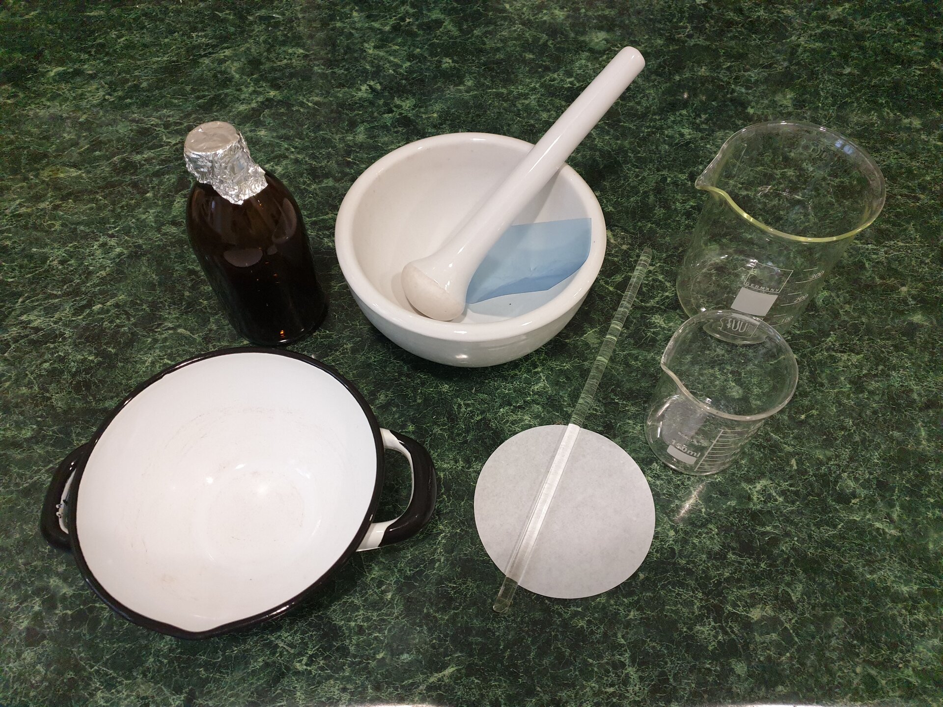 Zawiesiny. Rycina przedstawia szklaną butelkę z szyjką okrytą folią aluminiową, różnej wielkości szklane naczynia z podziałką, mające formę walca, a na górnej krawędzi wyżłobienie, które pomaga w przelewaniu. Ponadto na rycinie są przedstawione: miska, tłuczek (czyli moździerz z pistlem) oraz folia zabezpieczająca, okrągły, papierowy filtr, szklane mieszadełko i ceramiczna miska z uchami i wyżłobieniem znajdującym się na górnej krawędzi, ułatwiającym przelewanie. 