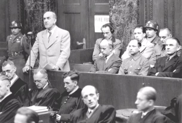 Zdjęcie przedstawia salę sądową. Przy kilku rzędach drewnianych ław siedzą mężczyźni. Jeden z nich stoi i przemawia do mikrofonu.