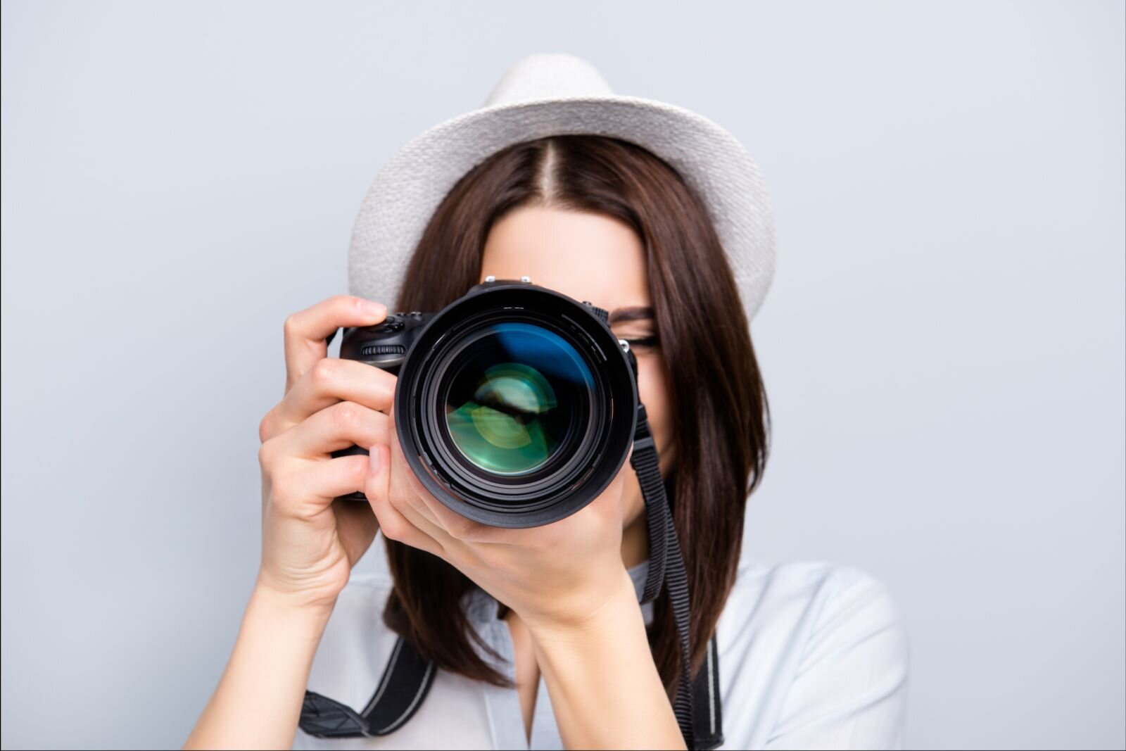 Zdjęcie przedstawia fotografującą dziewczynę. Jest zwrócona w naszą stronę, obiema rękami trzyma aparat na wysokości twarzy. Kapelusz na głowie i jasna koszula zlewają się z tłem. Ciemne włosy dziewczyny i obiektyw aparatu fotograficznego, który zasłania całą jej twarz są najmocniejszymi elementami zdjęcia. 