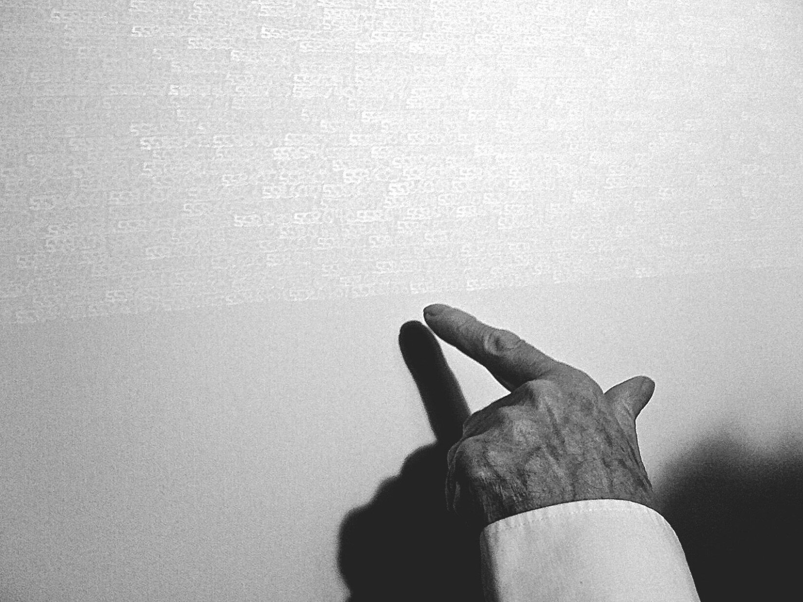 Fotografia przedstawia jasną powierzchnię, na której górnej części widoczne są rzędy białych wzorów, oraz dłoń wskazującą w ich kierunku. Dłoń znajduje się z prawej strony, na dole fotografii. Palec wskazujący skierowany jest ku rzędowi wzorów, a kciuk - w prawo. Pozostałe palce są zgięte. Na powierzchni dłoni widoczne są ciemniejsze żyły oraz zmarszczki. Widoczny jest fragment białego, koszulowego rękawa. Białe wzory na ścianie przypominają cyfry „5” umieszczone ściśle obok siebie, niektóre odwrócone. 