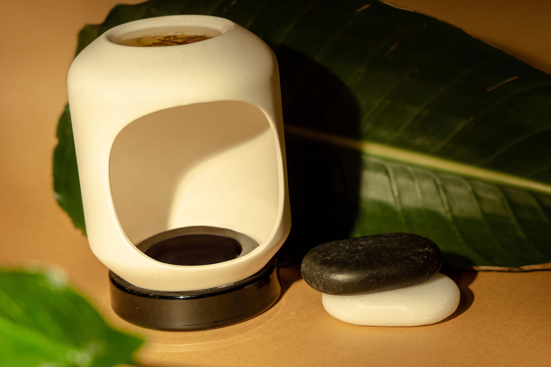 Grafika przedstawia obłe naczynie z wyciętym okrągłym otworem z przodu. Obok leżą dwa kamienie (czarny i biały) a za nimi duży liść. 