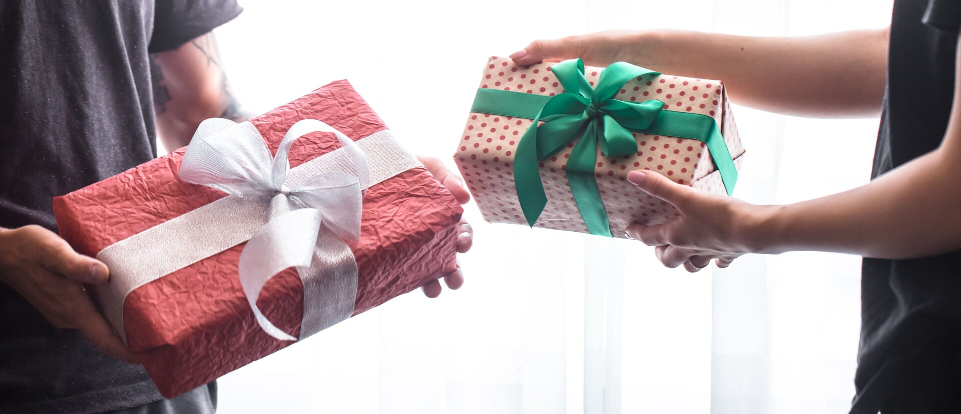 Zdjęcie przedstawia dwie postacie, widoczne od bioder do klatki piersiowej, stojące na przeciw siebie. Obydwoje trzymają w rękach prostokątne pudełka owinięte papierem prezentowym i przewiązane wstążką. Podają sobie wzajemnie prezenty. Człowiek stojący po prawo, ma mniejsze pudełko.