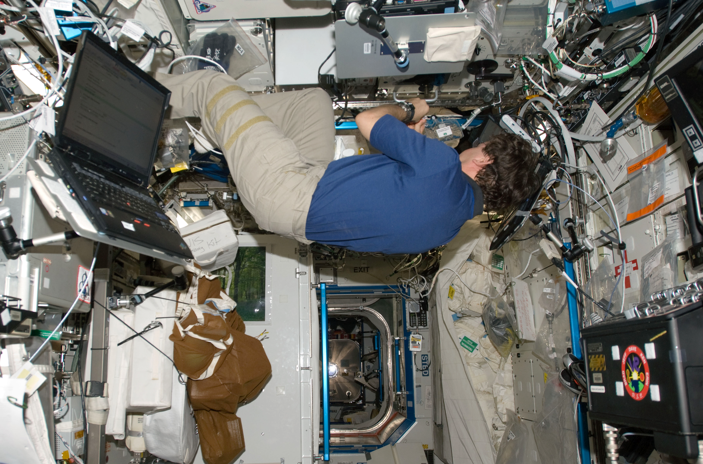 Fotografia wykonana z góry, ukazuje astronautę w stanie nieważkości w niewielkim pomieszczeniu na stacji kosmicznej. Astronauta widoczny ze swojego lewego boku manipuluje przy sprzęcie. Astronauta usytuowany jest na zdjęciu poziomo. Na ścianach pomieszczania przytwierdzone różne sprzęty oraz kable. Poniżej astronauty widoczne przejście do następnego pomieszczenia.
