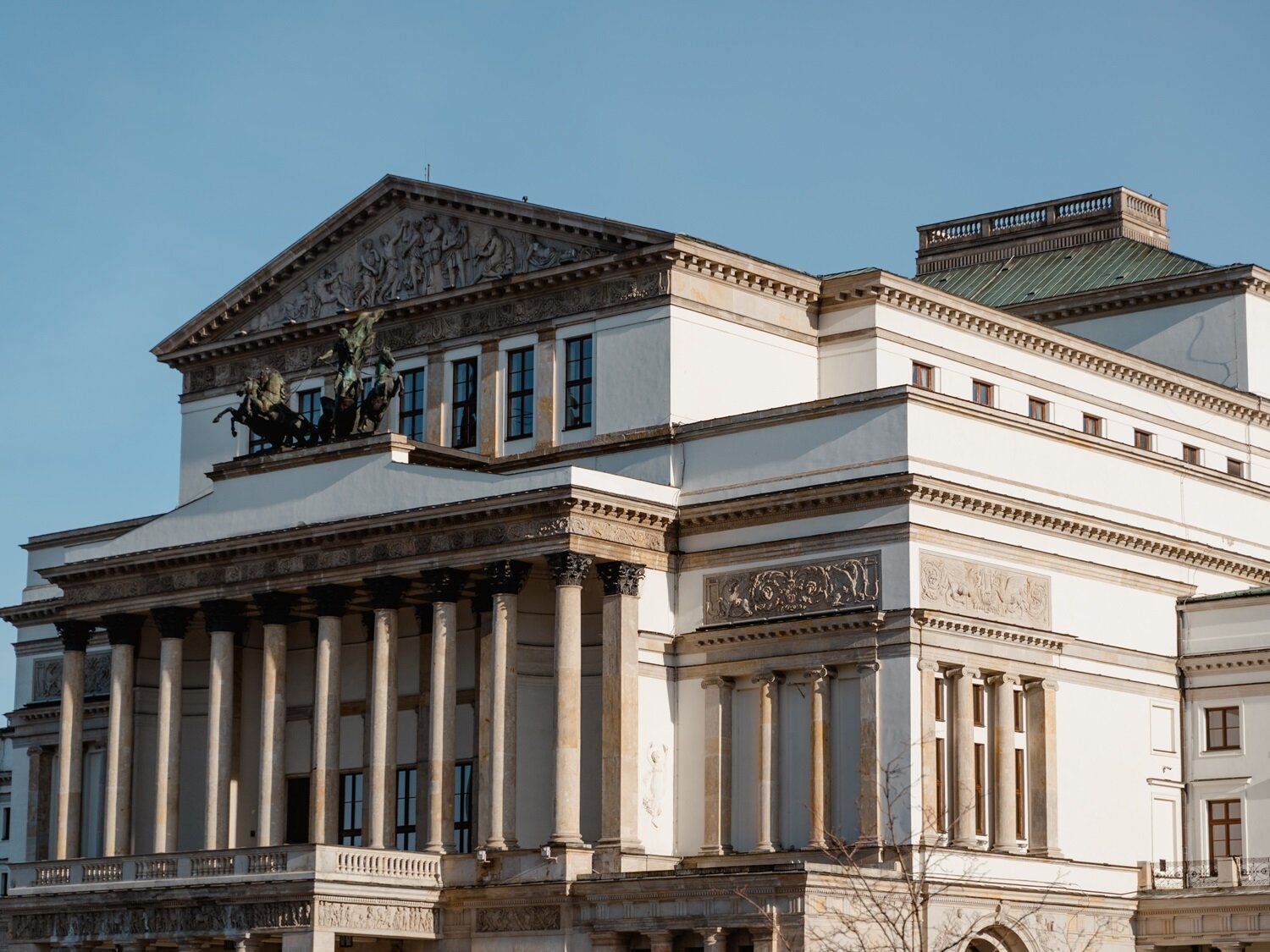 Zdjęcie przedstawia rozłożysty budynek zbudowany w stylu neoklasycznym, którego główna bryła ma kształt prostopadłościanu z fasadą zdobioną rzędem kolumn oraz półkolumn. Nad wejściem widoczna jest rzeźba przedstawiająca rydwan z końmi. Za rzeźbą widoczne jest piętro z oknami, zwieńczone trójkątnym tympanonem z płaskorzeźbą.