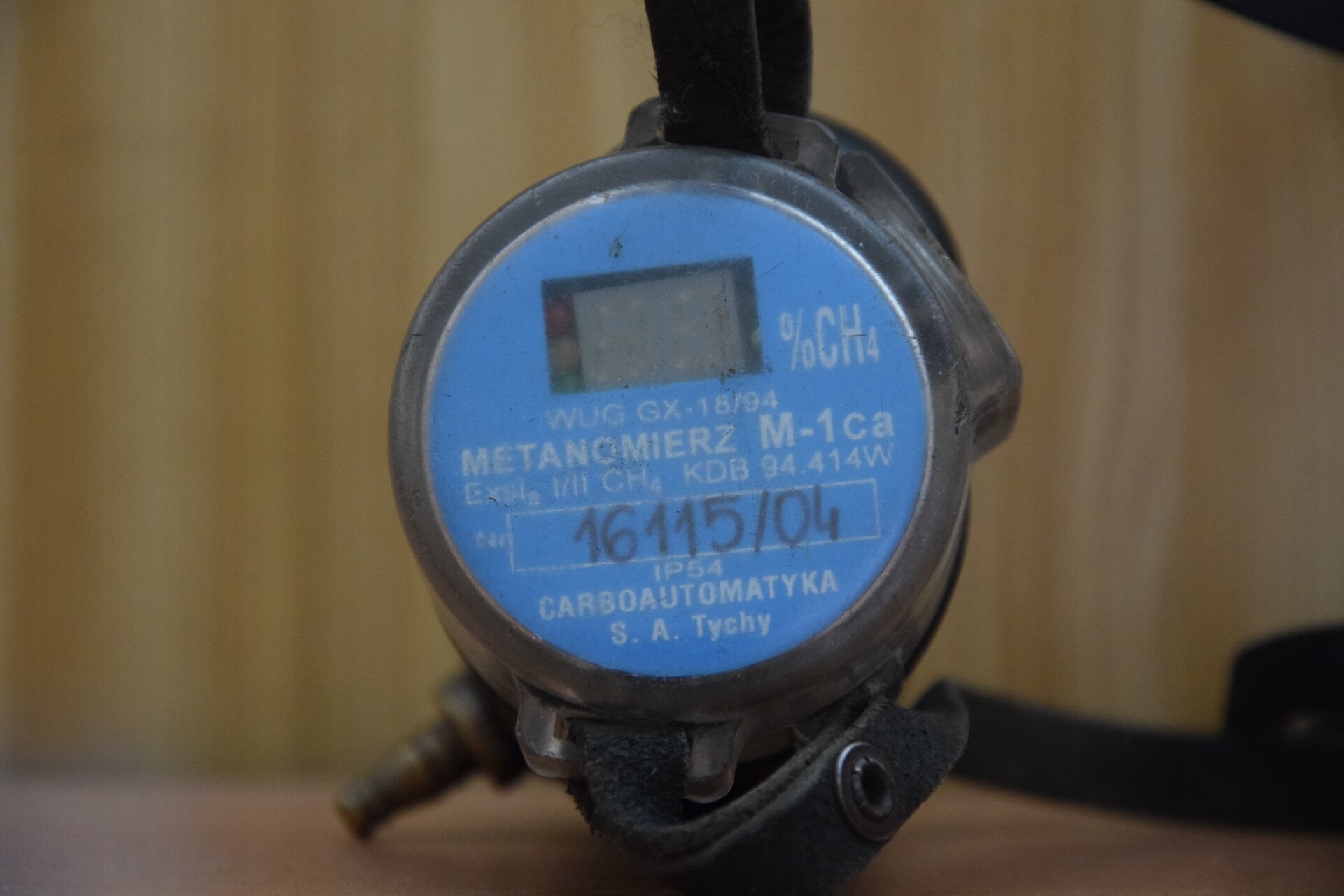 Na zdjęciu widoczny jest metanomierz. Górna podstawa metanomierza jest w kształcie okręgu. Na środku podstawy znajduje się niewielki prostokątny ekran. Obok ekranu widać oznaczenie: procent metanu. Poniżej umieszczony jest mapis: Metanomierz M jeden ca. Poniżej znajduje się numer seryjny: szesnaście tysięcy sto piętnaście łamane na zero cztery. Na dole metanomierza znajduje się nazwa firmy: Carboautomatyka spółka akcyjna Tychy. W tle widać fragmenty innych instalacji.Poniżej zdjęcia w prawym dolnym rogu widoczny jest przycisk odtwarzania. Tekst nagrania jest zgodny z tytułem zdjęcia.