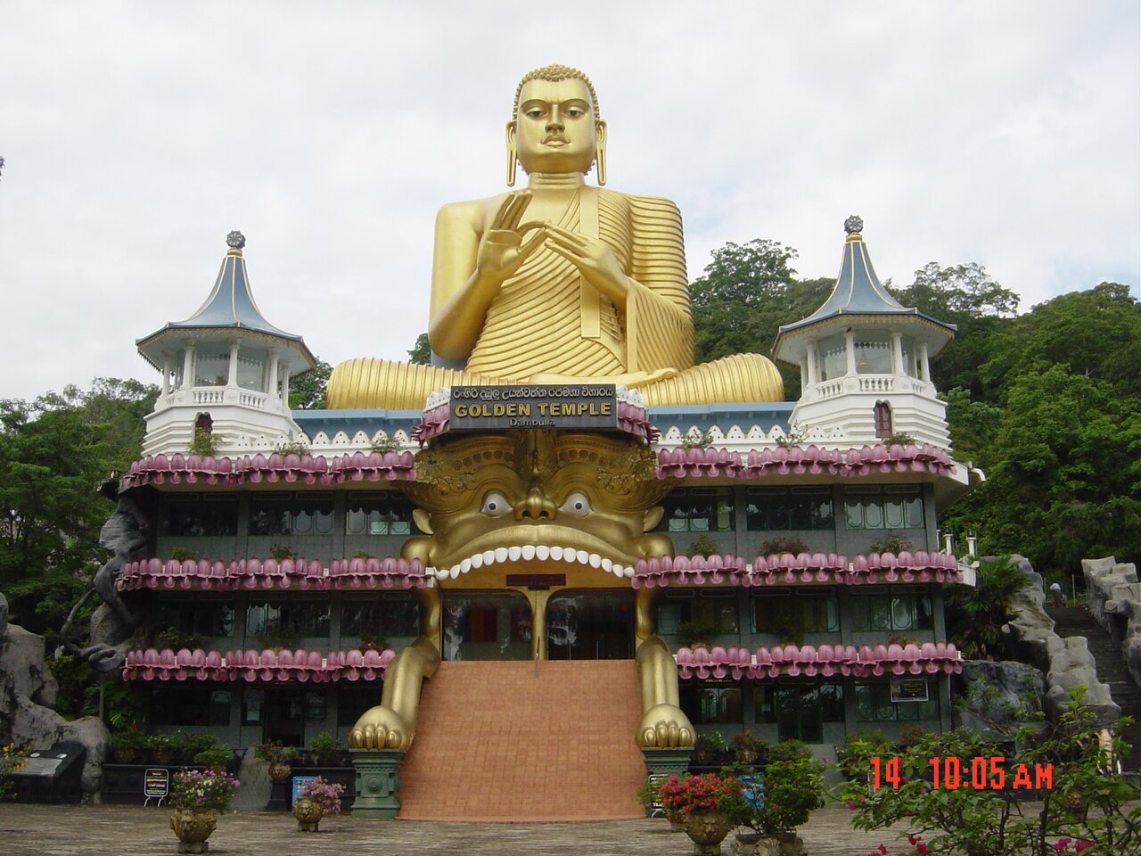 Kolorowe zdjęcie przedstawia świątynię hinduistyczną. Budynek ma trzy kondygnacje, każdą z nich zdobią różowe, półokrągłe wykończenia. Na szczycie świątyni widnieją dwie białe wieżyczki o niebieskich daszkach, pomiędzy którymi znajduje się olbrzymia, złota figura przedstawiająca Buddę siedzącego w pozycji kwiatu lotosu. Bóstwo ma krótkie włosy, prążkowaną szatę, długie ozdoby w uszach. Kciukiem i palcem wskazującym prawej dłoni przytrzymuje palce lewej dłoni. Poniżej widać tablicę z napisem "Golden Temple" [czyt. golden templ], która wisi nad wejściem, do którego prowadzą strome schody o poręczach przypominających łapy zwierzęcia, zwieńczonego daszkiem w postaci złotej głowy stwora (widoczne oczy, nos i górny rząd białych zębów). Przed świątynią stoją donice z kwiatami. Za nią rosną drzewa. Niebo jest błękitne.