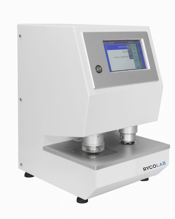 Zdjęcie przedstawia miernik gładkości metodą Bendsena, w ujęciu z ukosa. Widoczne jest urządzenie w kształcie zbliżonym do prostopadłościanu, ze ściętą pod kątem górną częścią przedniej ściany oraz głębokim podcięciem w dolnej części, które tworzy rodzaj blatu. Na ściętej ściance urządzenia widoczny jest wyświetlacz w srebrnej ramie, po lewej stronie której widoczny jest okrągły przycisk. W podcięciu widoczne są dwa cylindryczne elementy dotykające srebrnego blatu. Urządzenie stoi na czterech czarnych, niskich nóżkach, z których widoczne są tylko trzy. Po prawej stronie podstawy widoczny napis Rycolab.