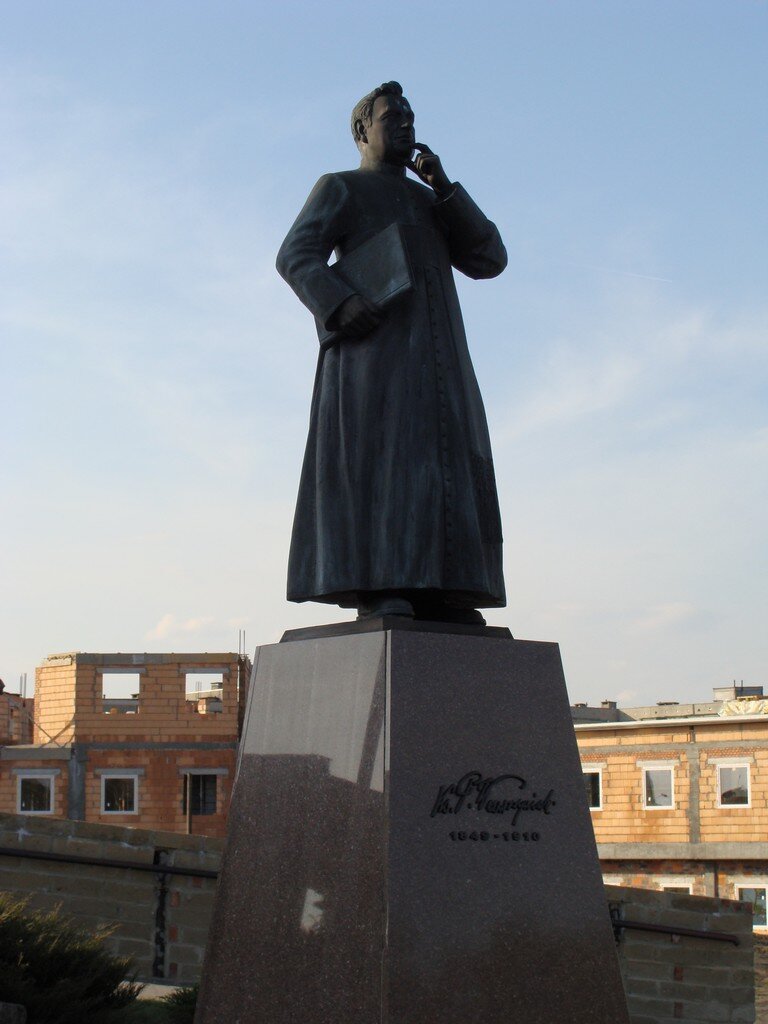 Na zdjęciu znajduje się pomnik księdza Piotra Wawrzyniaka ustawiony na wysokim kamiennym podeście. Jest on ubrany w szaty księdza. Ma krótkie włosy. W prawej ręce trzyma dużą książkę, a lewą trzyma się na podbródek. 