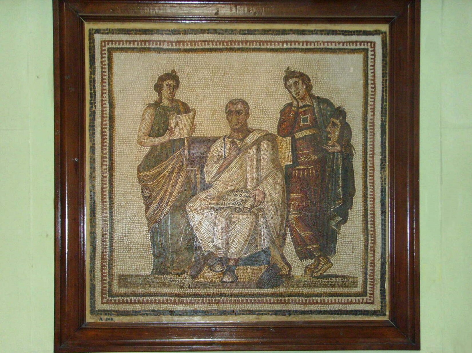 Na zdjęciu znajduje się mozaika „Wergiliusza z manuskryptem Eneidy, otoczonego przez Muzy: Klio i Melpomenę”. Mozaika znajduje się w drewnianej, brązowej ramie na jasnozielonym tle. Na środku siedzi mężczyzna w długiej, jasnej szacie i jasnych butach. Na kolanach trzyma zwój papirusu. Z prawej strony stoi postać w czerwonej szacie ze złotymi zdobieniami i zielonym materiałem na lewym ramieniu. W lewej dłoni trzyma maskę, a prawą dotyka swojego policzka. Z lewej strony mężczyzny stoi druga postać. Ma długą, zielono‑złotą szatę. W dłoniach trzyma zwój papieru. Tło mozaiki jest jasne.