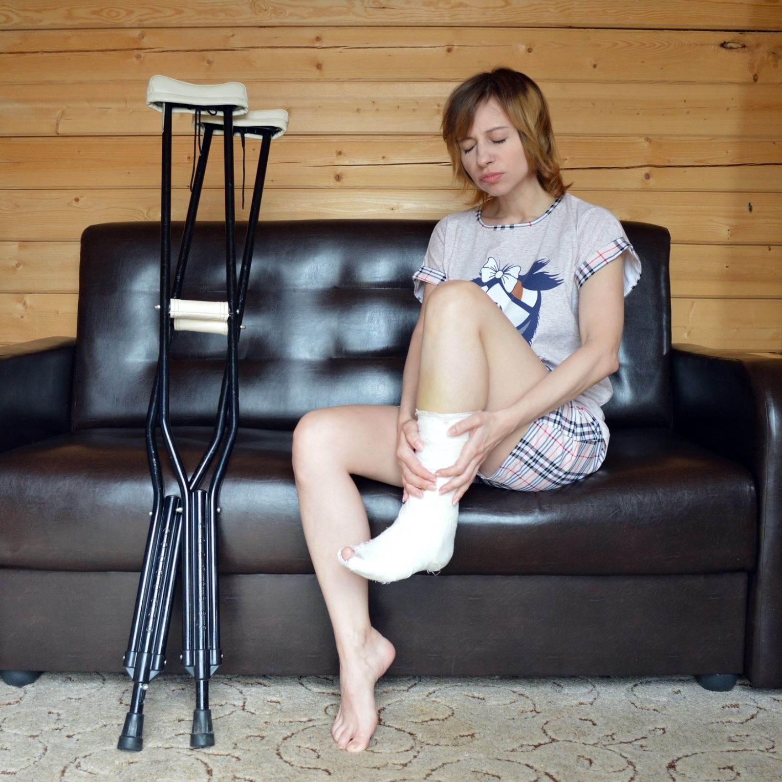 Zdjęcie przedstawia młodą kobietę, siedzącą na dużej czarnej, skórzanej kanapie. Lewą nogę ma podniesioną do góry, zgiętą w kolanie. Jej lewa stopa i dolna część łydki jest w gipsie. Prawa noga jest zgięta, stopa lekko podniesiona i oparta na palcach. Kobieta ubrana jest w pidżamę złożoną z koszulki z krótkim rękawem i szortów. Na lewo od niej, oparte o kanapę, stoją dwie kule ortopedyczne. W tle widoczna ściana z drewnianych desek, a na podłodze szary dywan.