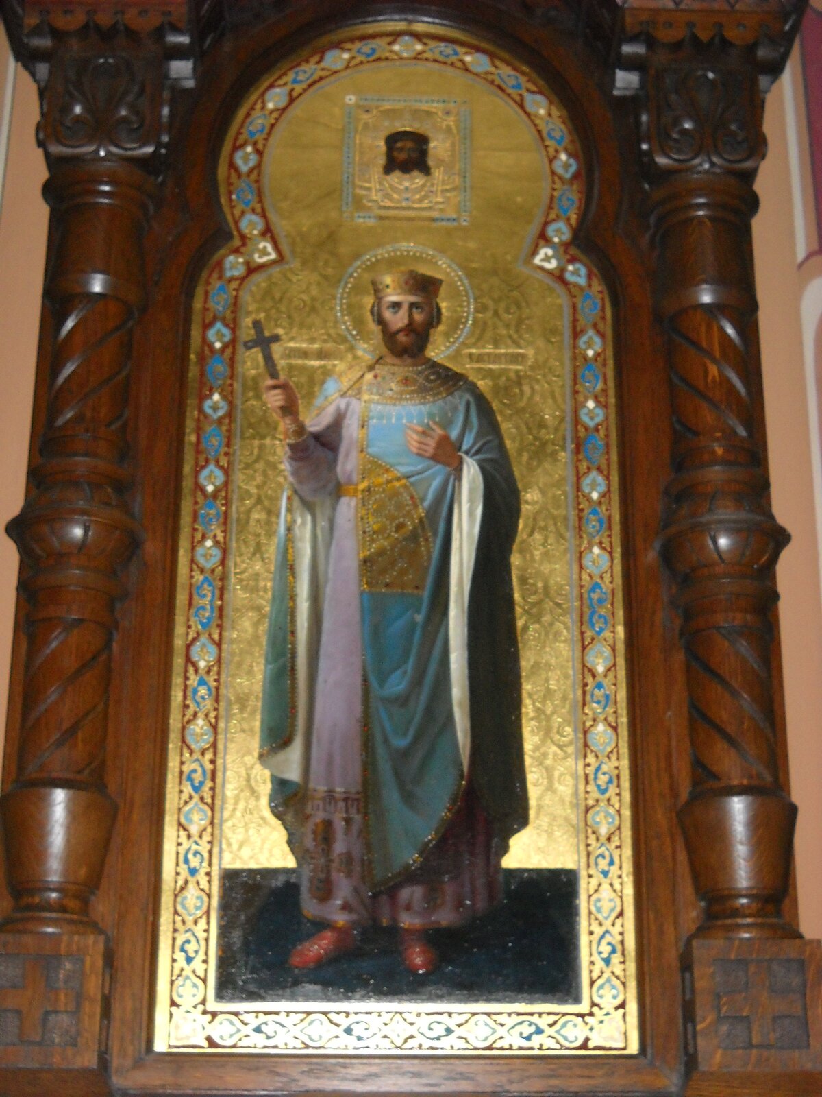 Fotografia przedstawia widok na ikonę św. Konstantyna, która znajduje się w cerkwi pod wezwaniem Wszystkich Świętych w Piotrkowie Trybunalskim. Ikona przedstawia postać św. Konstantyna, który został ukazany jako mężczyzna w średnim wieku o średniej długości zaroście, ubrany w biało‑niebieską szatę sięgającą do kostek, na której obecne są złote zdobienia. Mężczyzna jedną dłoń trzyma na klatce piersiowej, na wysokości serca, a w drugiej dłoni ma krzyż. Portret został umieszczony na złotym tle, w ozdobnej ramce. Powyżej widoczna jest podobizna mężczyzny o długich włosach oraz krótszym zaroście. Po bokach widoczne są drewniane kolumny ścienne. 