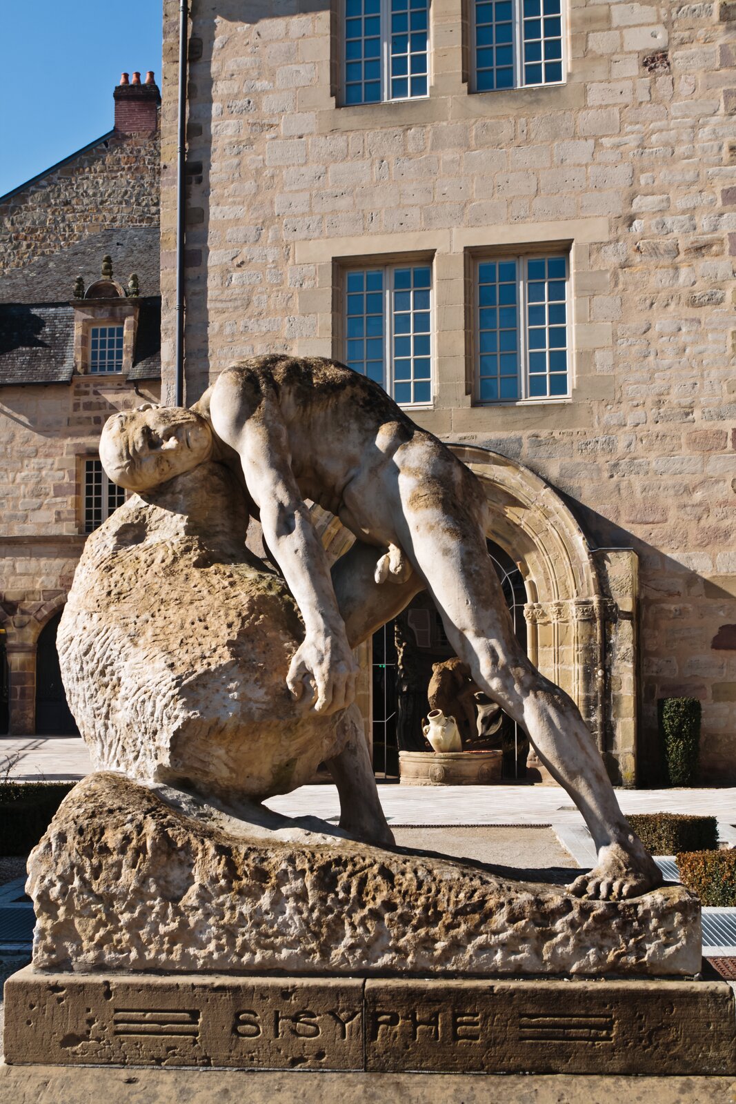 Ilustracja przedstawia rzeźbę Syzyfa, który z wielkim mozołem toczy kamień. Głowę ma położoną na kamieniu i próbuje go od dołu złapać, aby potoczyć go dalej. Mężczyzna jest nagi, ma napięte mięśnie, zmęczoną twarz i zgarbione plecy. Rzeźba stoi przed budynkiem z piaskowca z prostokątnymi oknami. Niebo jest niebieskie, świeci słońce.