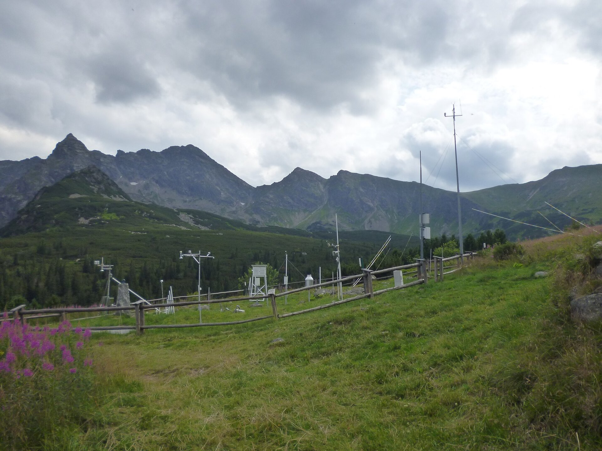 Zdjęcie przedstawia stację meteorologiczną znajdującą się na Hali Gąsienicowej, powyżej schroniska na Murowańcu w Tatrach. Widać stok porośnięty trawą. Na środku widać otoczoną płotem stację. Widoczne są w niej liczne narzędzia pomiarowe o różnych kształtach. W tle widać wysokie szczyty.