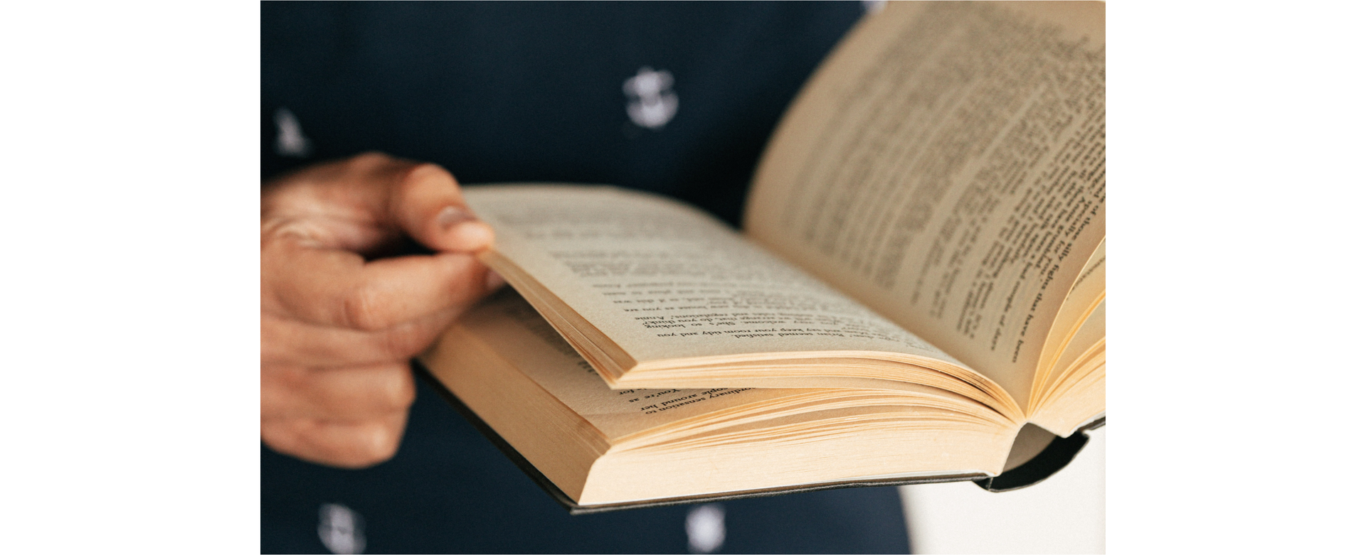 Zdjęcie przedstawia zbliżenie na otwartą książkę. Widoczna jest dłoń osoby trzymającej książkę.