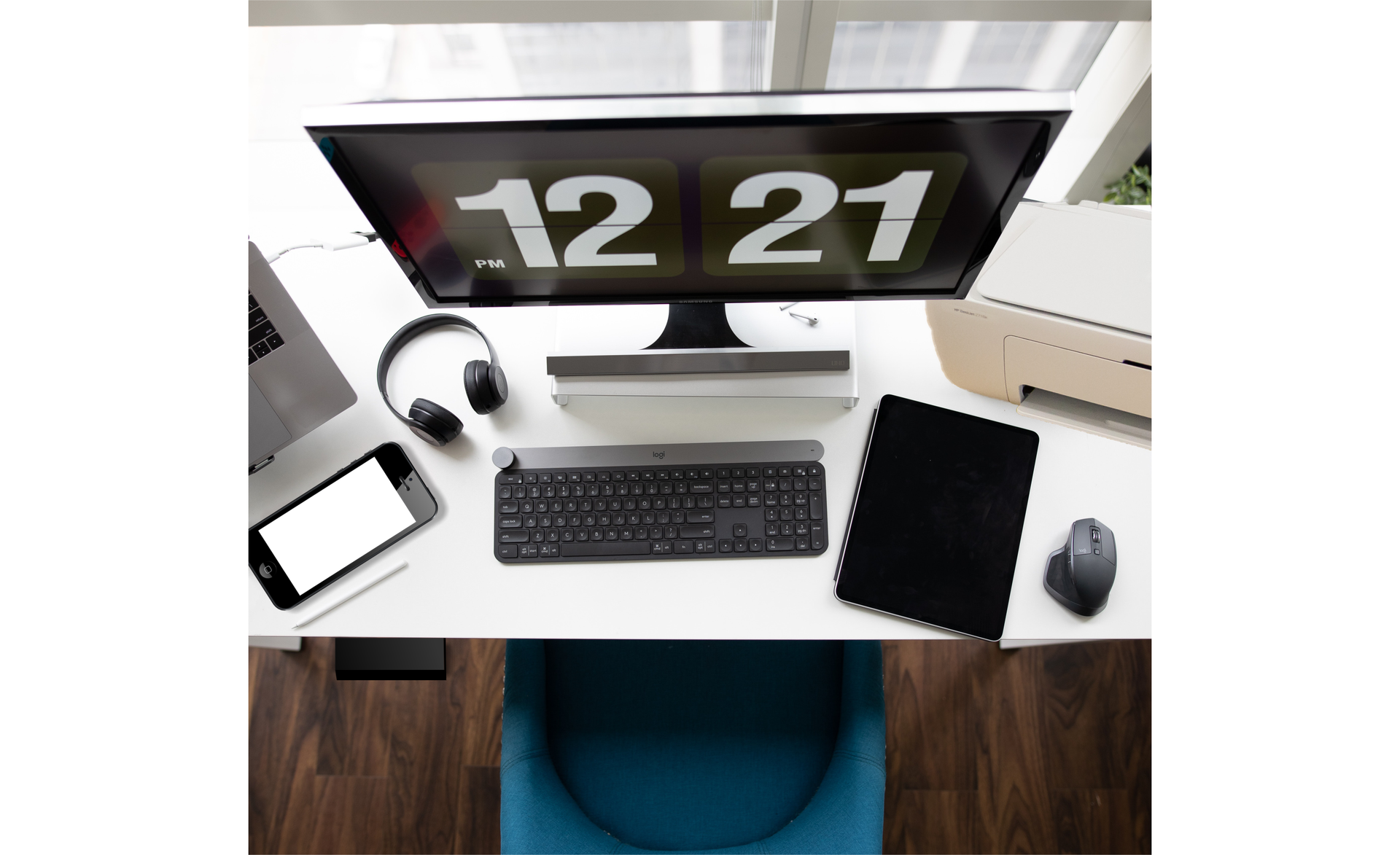 Zdjęcie przedstawia miejsce w pracy w biurze. Biurko stoi przy oknie, na środku stoi monitor, a przed nim leży klawiatura. Po prawej stronie monitora znajdują się: tablet, myszka i drukarka. Po lewej stronie monitora leżą słuchawki i telefon.