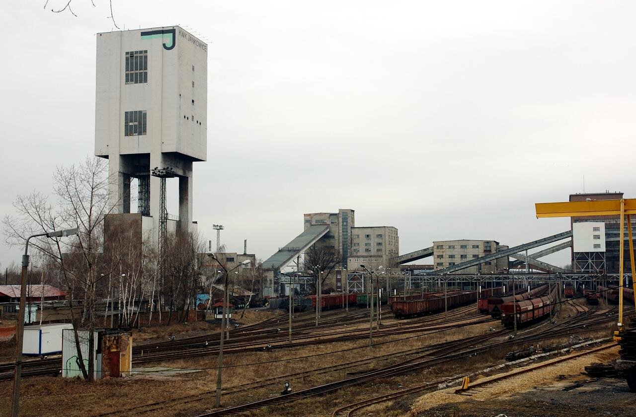 Zdjęcie przedstawia powierzchnię kopalni z szybem. Szyb jest prostokątny, znajduje się na wysokich podporach. Na pierwszym planie jest torowisko. W tle są budynki połączone z taśmociągami.
