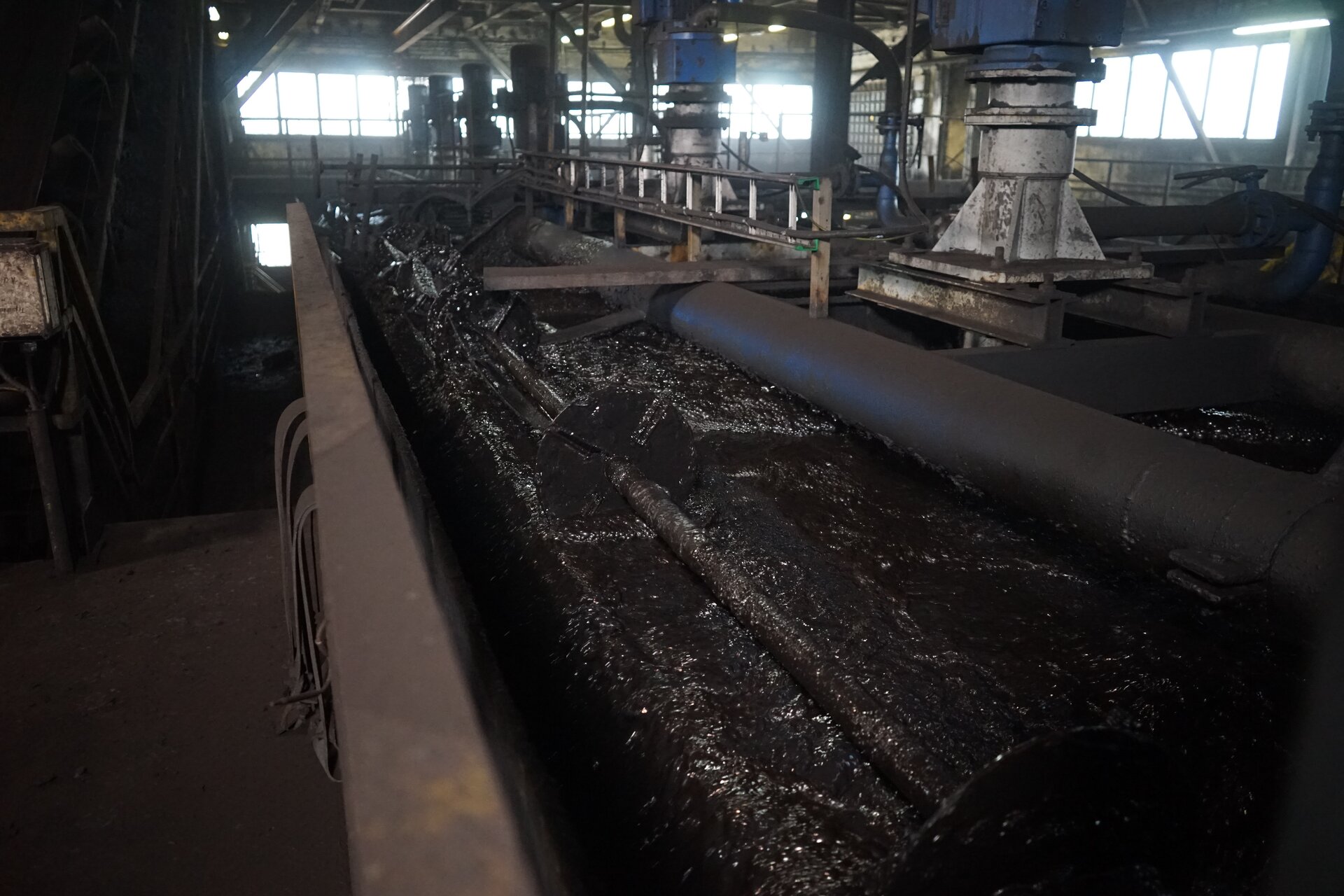 Zdjęcie przedstawia podzespoły połączone komorami pośredniczącymi. Razem składają się one na flotownik. Po prawej stronie stronie zbiorniki. w których znajdują się komory robocze wypełnione czarnymi mętami flotacyjnymi. W ich wnętrzach zanurzone są pionowe wały. Nad zbiornikami zamontowane są silniki z zaczepionymi pasami napędowymi. Wprawią one w ruch zabudowane na dnie komór roboczych wirniki. Po prawej stronie powyżej poziomu lustra cieczy zabudowane jest koryto odbiorcze. Przed nim znajdują się zgarniacze skrzydełkowe. Są one częściowo zanurzone w pianie flotacyjnej. Poniżej zdjęcia znajduje się przycisk multimedialny z nagraniem audio. Treść nagrania jest zgodna z tytułem zdjęcia.