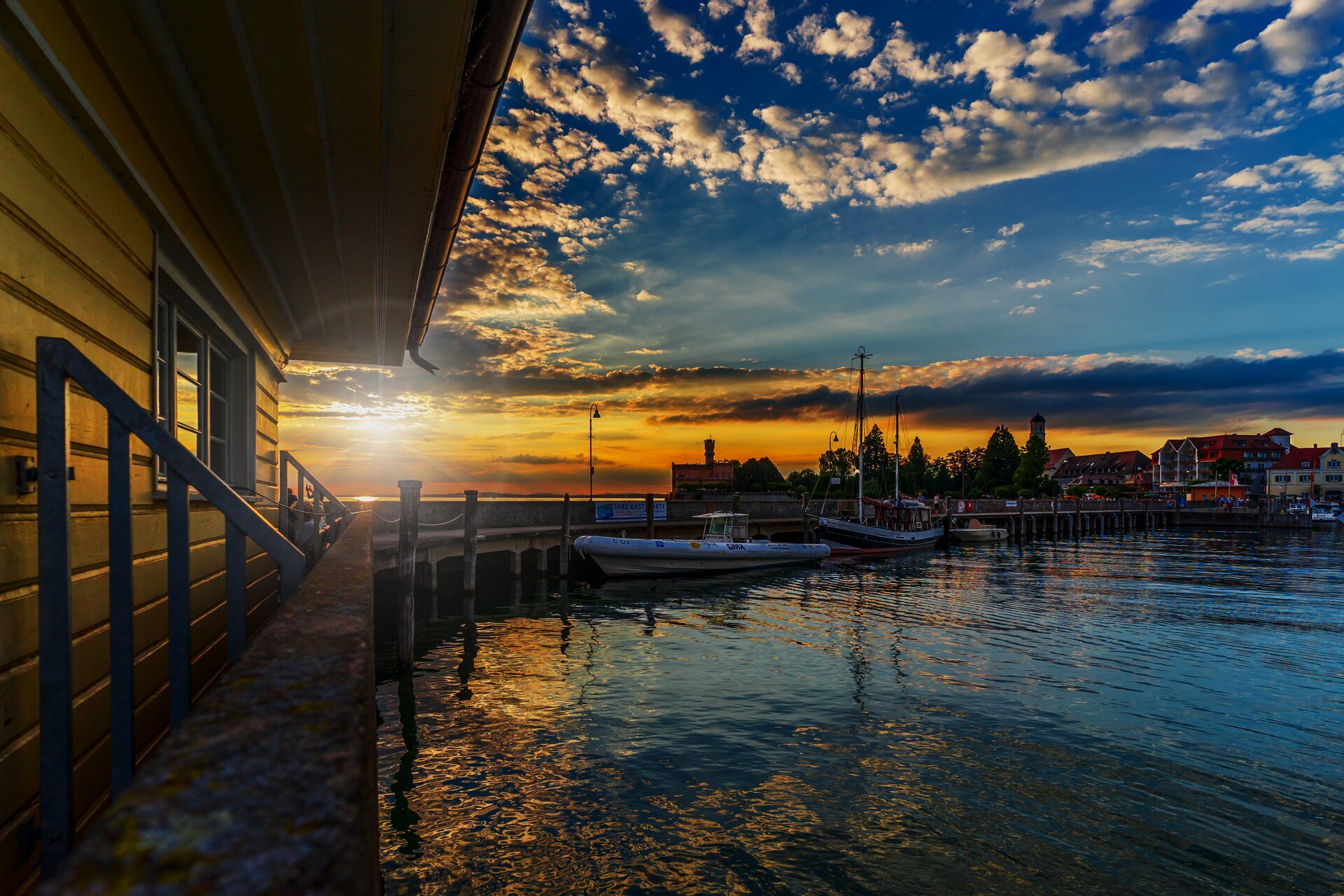 Ilustracja przedstawia widok na jezioro o zachodzie słońca. Z przodu ilustracji znajduje się drewniany budynek z pomostem. Z tyłu za jeziorem widać kontury miasta.