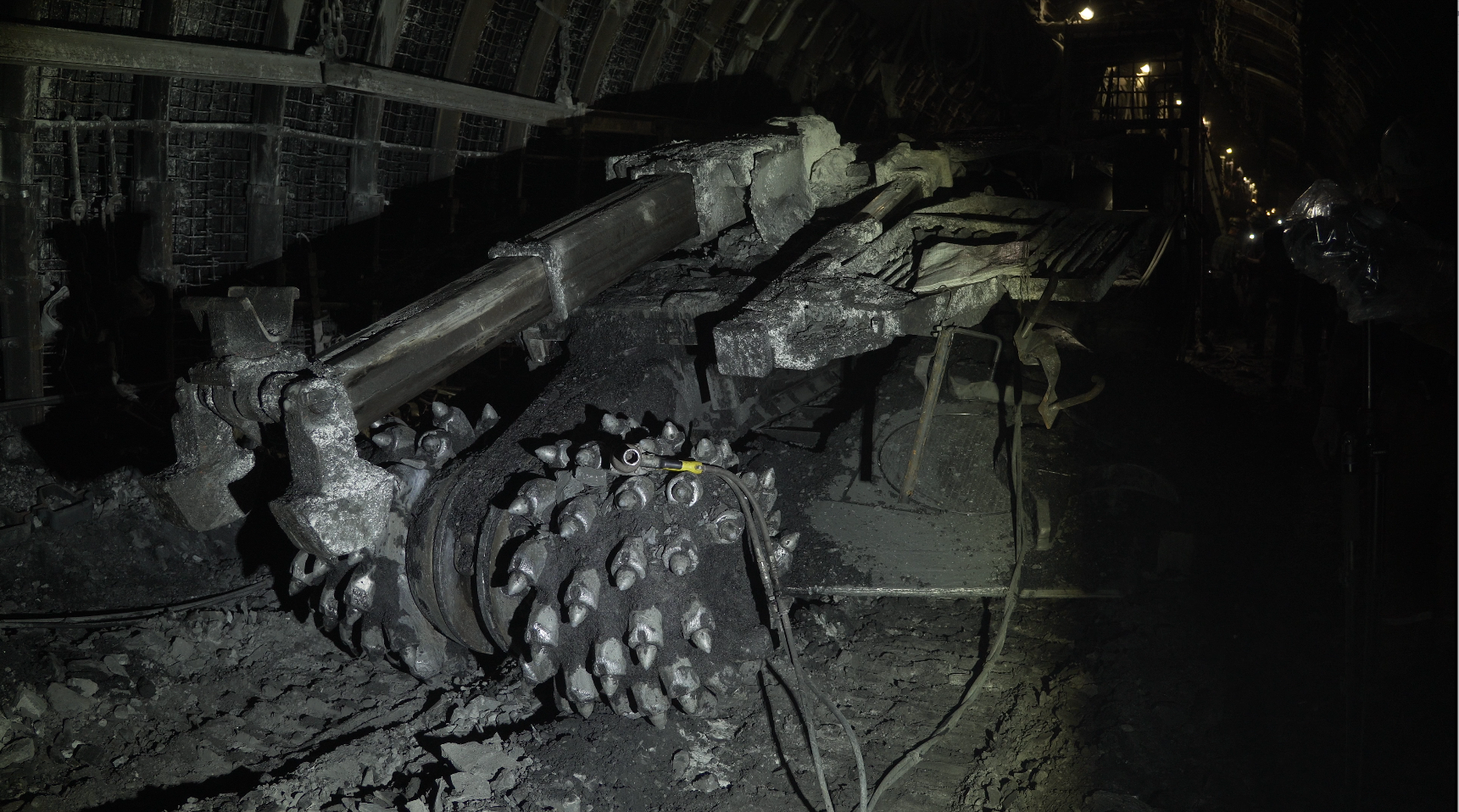 Na zdjęciu widoczny jest kombajn chodnikowy przystosowany do podnoszenia stropnicy obudowy stalowej podatnej. Na zdjęciu widoczna jest przednia część kombajnu. Od góry obudowy odchodzi teleskopowe ramię o kształcie prostopadłościanu. Ramię zakończone jest chwytakiem o kształcie litery U. Poniżej ramienia znajduje się drugie ramie z organem roboczym. Organ roboczy stanowią dwie głowice o kształcie półkul. Na powierzchni każdej z półkul znajdują się stożkowe zęby. Na prawej półkuli położony jest klucz przypominający pistolet.W tle widać słabo oświetlone wyrobisko korytarzowe.