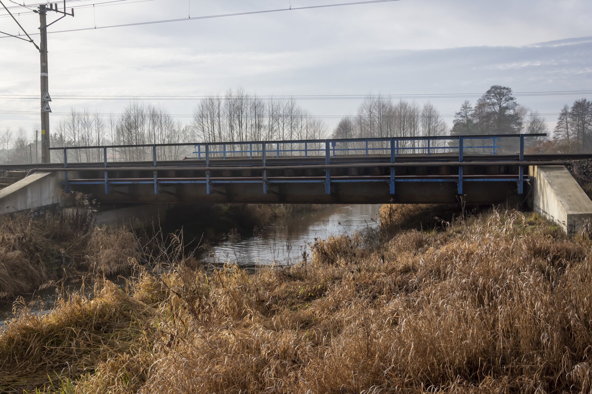 Zdjęcie przedstawia most belkowy. Posiada on jedno przęsło, które opiera się jedynie o podpory skrajne, tak zwane przyczółki. Most nie jest zbyt wysoki, rozciąga się nad rzeką. Na moście znajduje się niebieska barierka. Obok mostu znajduje się słup elektryczny. Widoczne są również w tle drzewa i niebo.