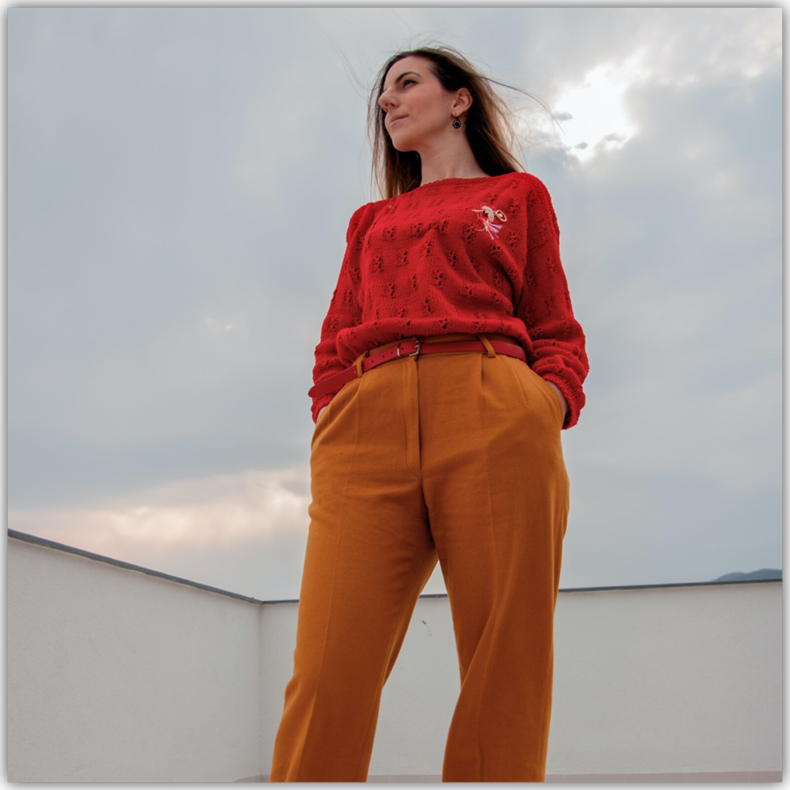 Zdjęcie przedstawia kobietę stojącą na dachu budynku. W tle niebo. Kobieta ma długie brązowe włosy, ma na sobie czerwony sweter i pomarańczowe spodnie. Ręce trzyma w kieszeniach.