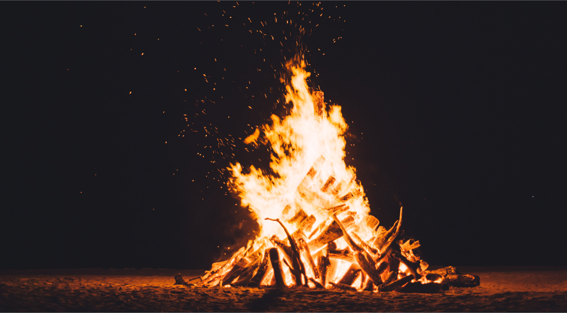 Duże ognisko nocą. Płomienie unoszone są wysoko do góry. Dookoła rozchodzą się iskry.