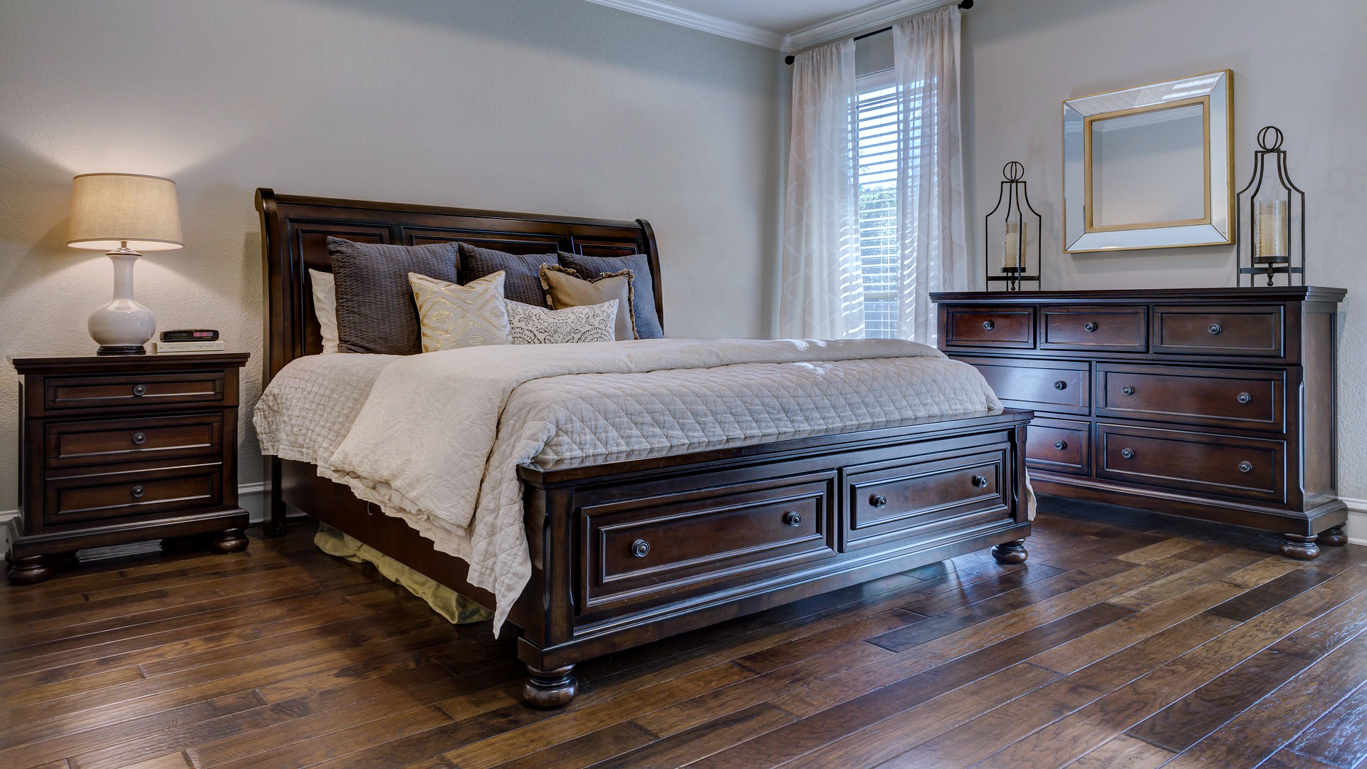 Zdjęcie przedstawia dwuosobowe łóżko w sypialni. Po lewej stronie stoi stolik nocny z lampką. Po prawej stronie łóżka znajduje się ściana z oknem, obok którego stoi komoda.