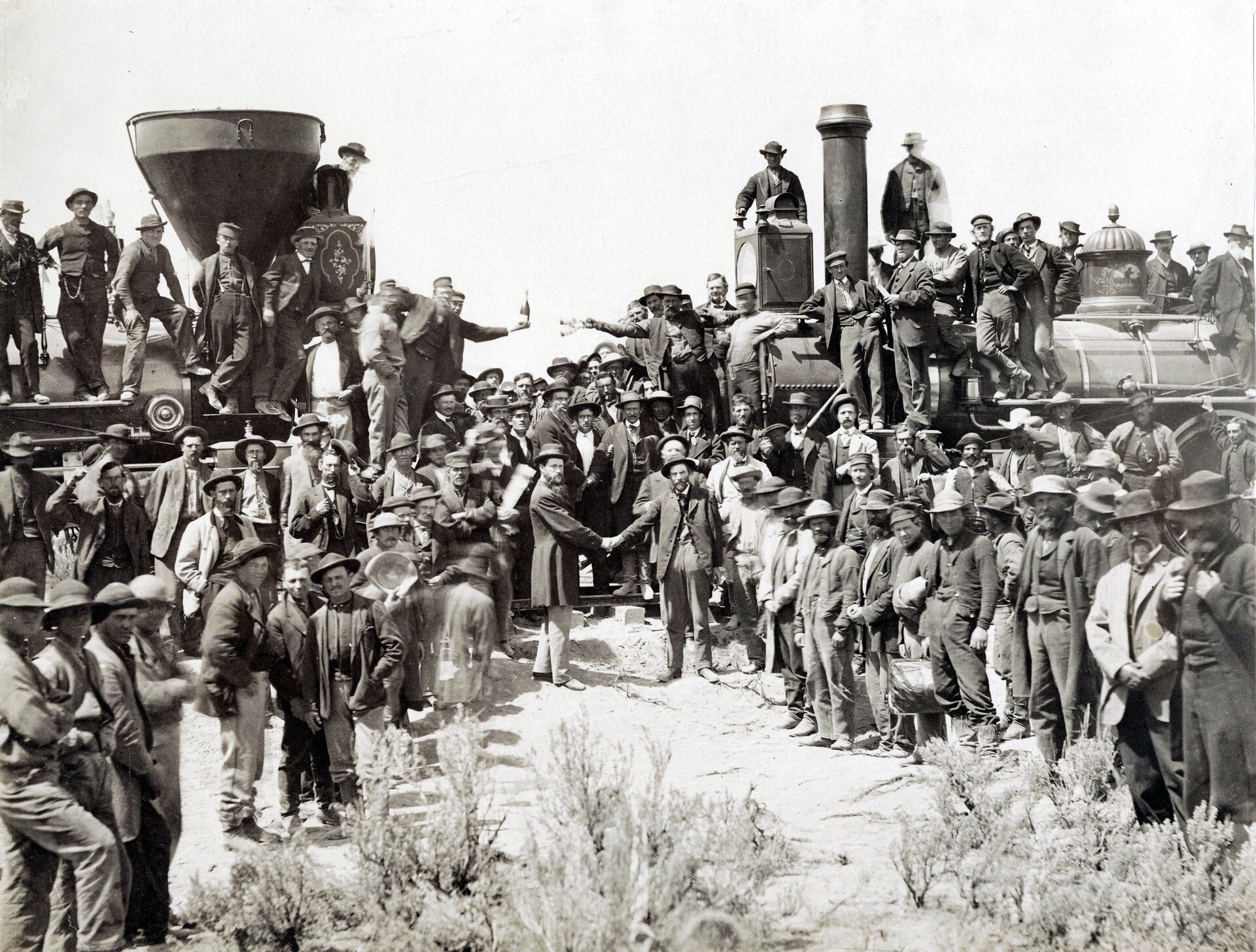 Zdjęcie przedstawia grupę ludzi stojących przed dwoma ustawionymi do siebie przodem lokomotywami. Część z robotników siedzi na maszynach, część stoi przed nimi. Na pierwszym planie dwóch mężczyzn ubranych elegacko, podających sobie ręce.