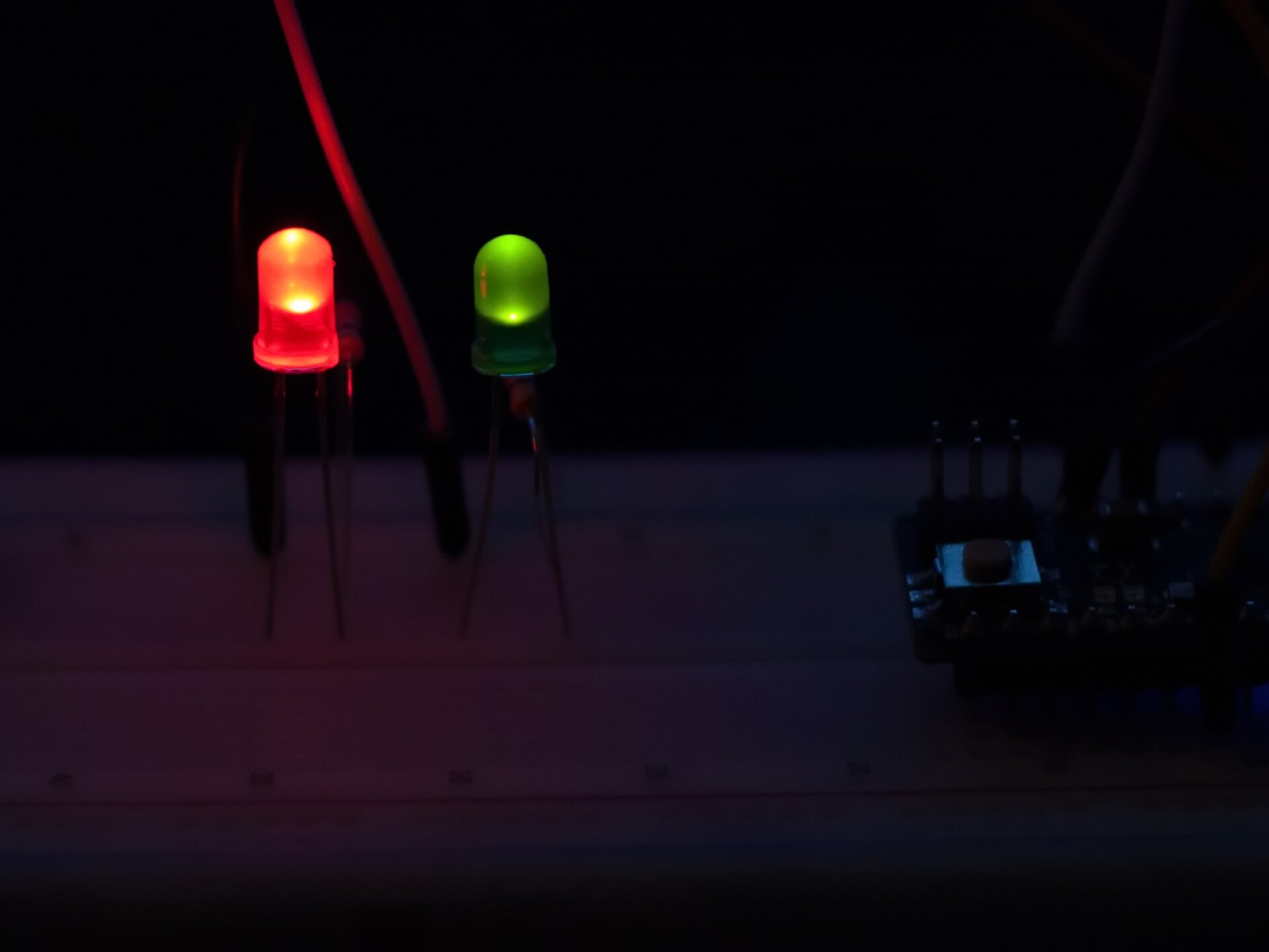 Rys. a. Na zdjęciu widoczne są dwie diody elektroluminescencyjne LED podłączone do prądu. Jedna z nich świeci na czerwono a obok niej znajduje się druga świecąca na zielono. Po prawej stronie widać zacieniony sterownik służący do włączania i wyłączania obu elementów.
