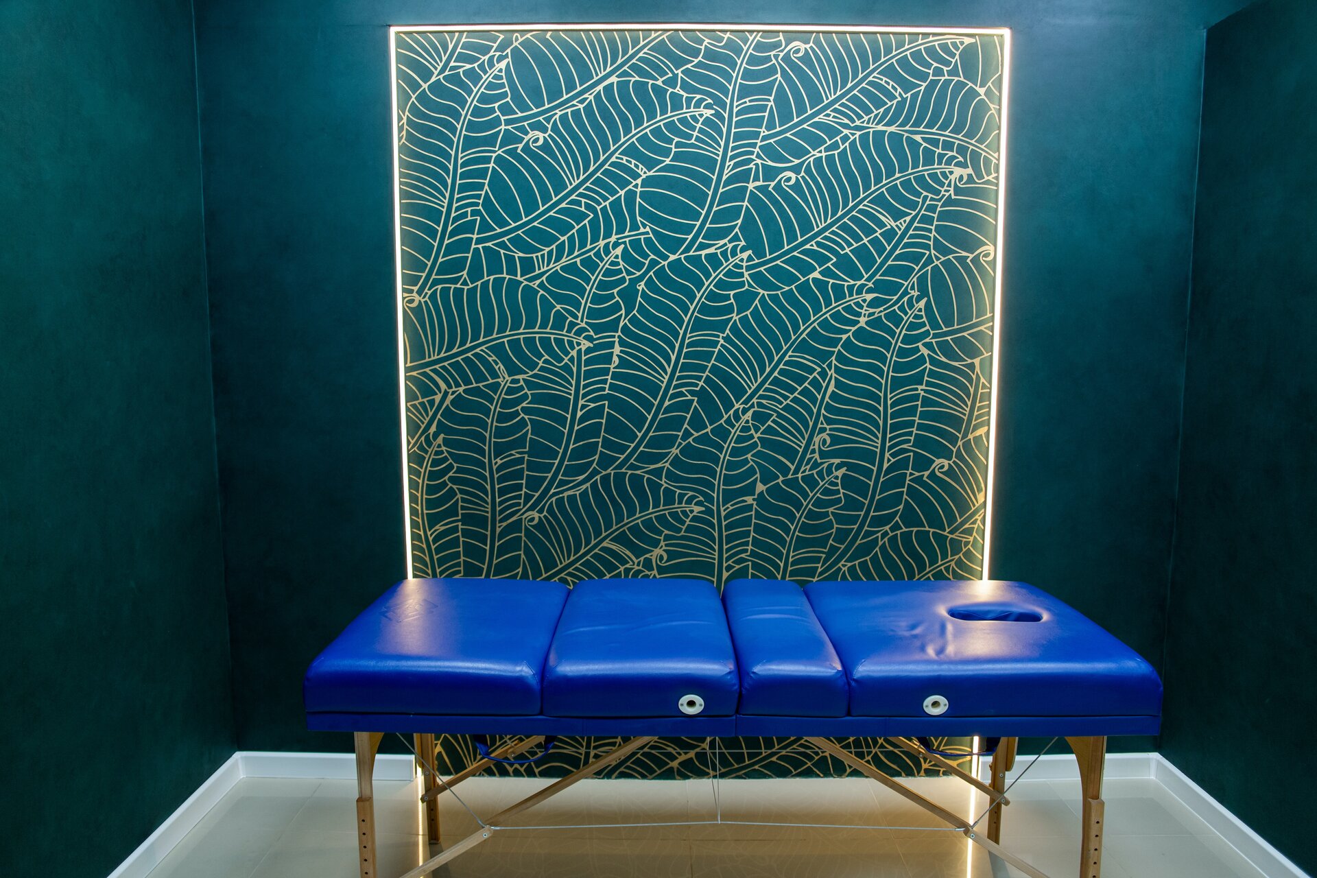 Zdjęcie przedstawia tapicerowany stół do masażu. Stół znajduje się w niewielkim pomieszczeniu z dekoracyjną ścianą. Stół składa się z kilku prostokątnych części różnej wielkości ściśle do siebie przylegających. Na jednym końcu stołu znajduje się otwór. Stół obito niebieską ekoskórą.