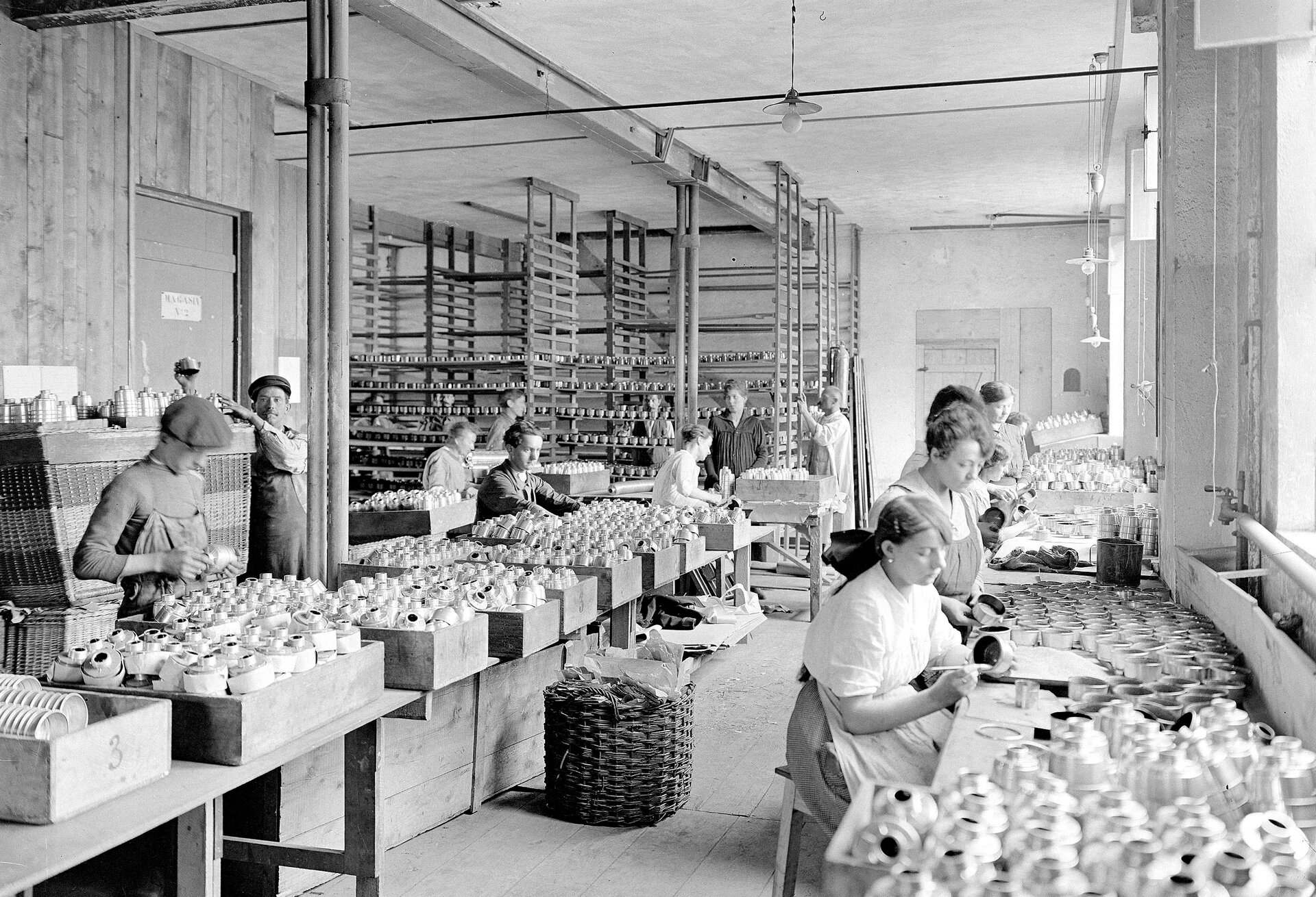 Zdjęcie przedstawia pomieszczenie fabryki. Przy stołach siedzą kobiety w prostych sukniach. Składają i malują elementu do masek gazowych, które umieszczone są w drewnianych pudełkach. W tle stoją półki z elementami.