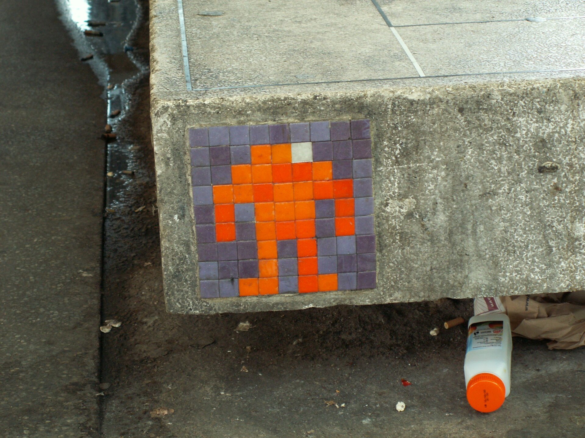 Ilustracja przedstawia wlepkę ukazującą symbol ludzika złożonego z pomarańczowych kwadracików na szarym tle. Umieszczona jest na betonowej płycie.