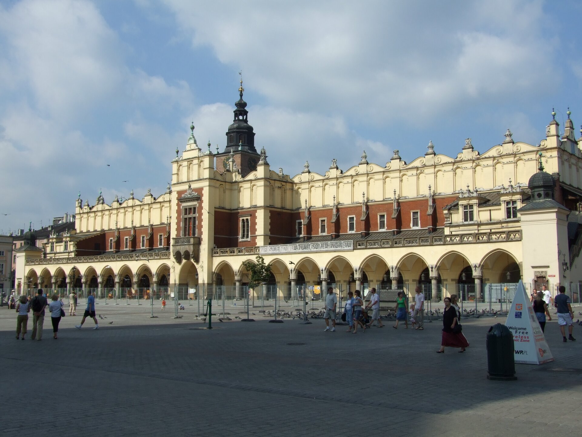 Na zdjęciu długi dwupiętrowy budynek. W dolnej kondygnacji znajdują się pomieszczenia otwarte na zewnątrz, ograniczone filarami wzdłuż lica muru, pokryte szarym dachem. Kolor elewacji w dolnej kondygnacji jest jasnożółty. Druga kondygnacja jest zamknięta, w ścianie znajdują się liczne okna. Elewacja w kolorze czerwonym. U góry budynku attyka - piękne zdobienia osłaniające dach.  W tle widoczna wieża budynku stojącego z tyłu. 