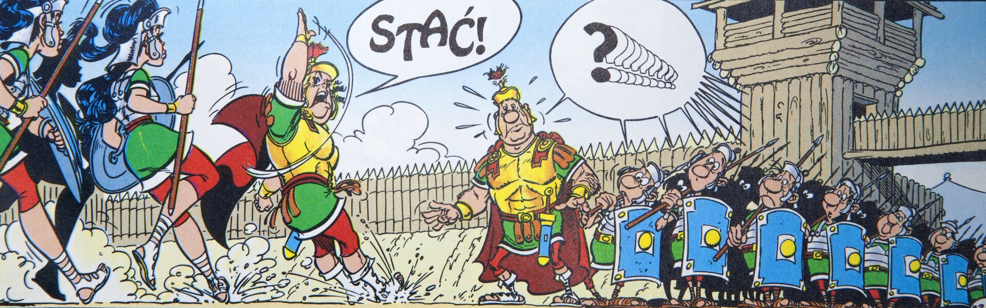 Ilustracja przedstawia fragment komiksu „Asterix. Róża i miecz”. Ukazuje dwa oddziały wojowników z dowódcami na czele. Żołnierze idą ku sobie w szyku. Dowódca lewego oddziału podnosi wyprostowaną rękę. Od niego unosi się dymek ze słowem: Stać!. Nad prawym oddziałem oraz jego dowódcą unosi się dymek ze zwielokrotnionym znakiem zapytania. W tle widoczna jest drewniany mur obronny z bramą i wierzą strażniczą. Nad nim rozciąga się błękitne niebo z chmurami.