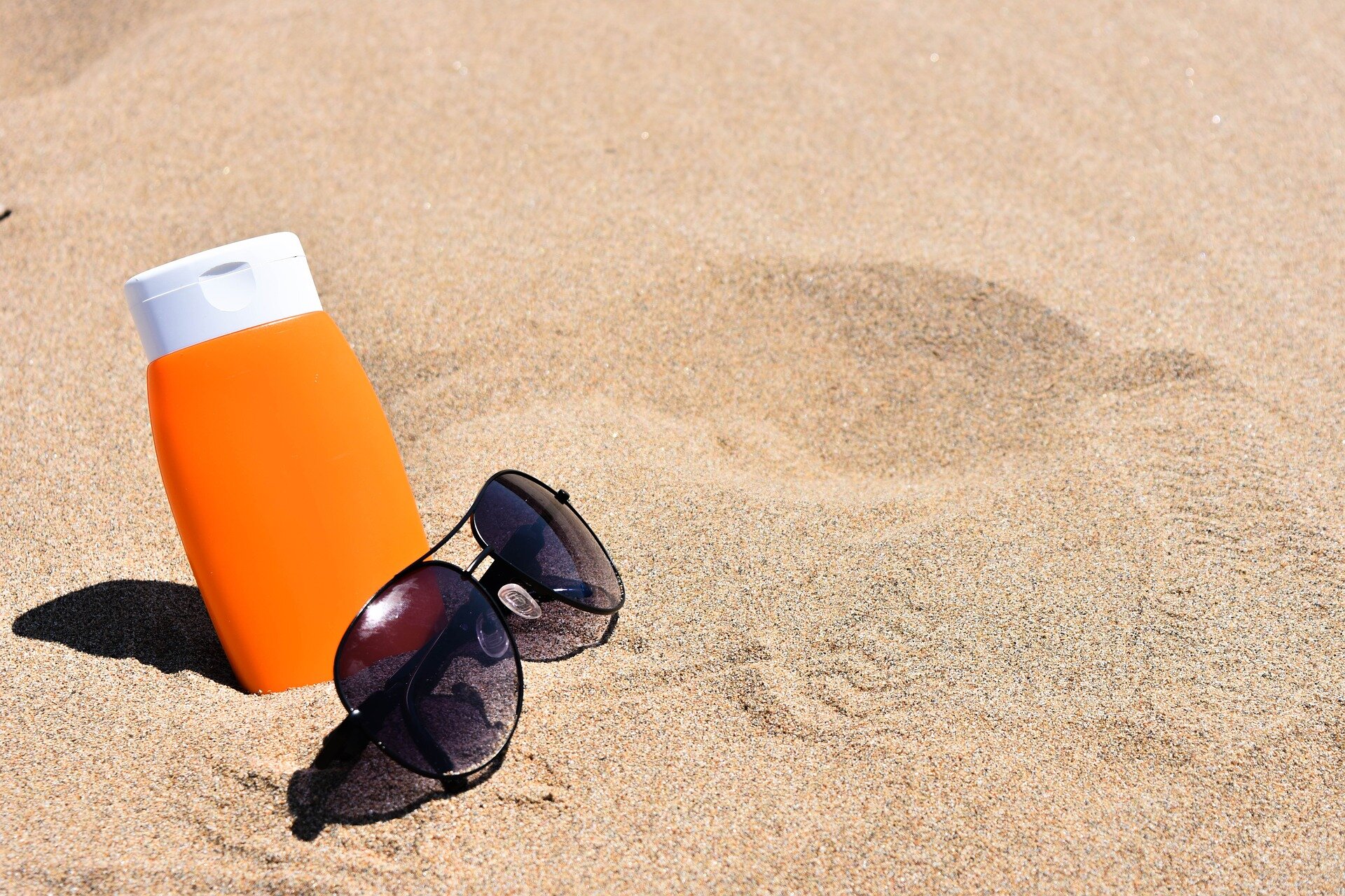Zdjęcie przedstawiające krem do opalania oraz okulary przeciwsłoneczne leżące na piasku.