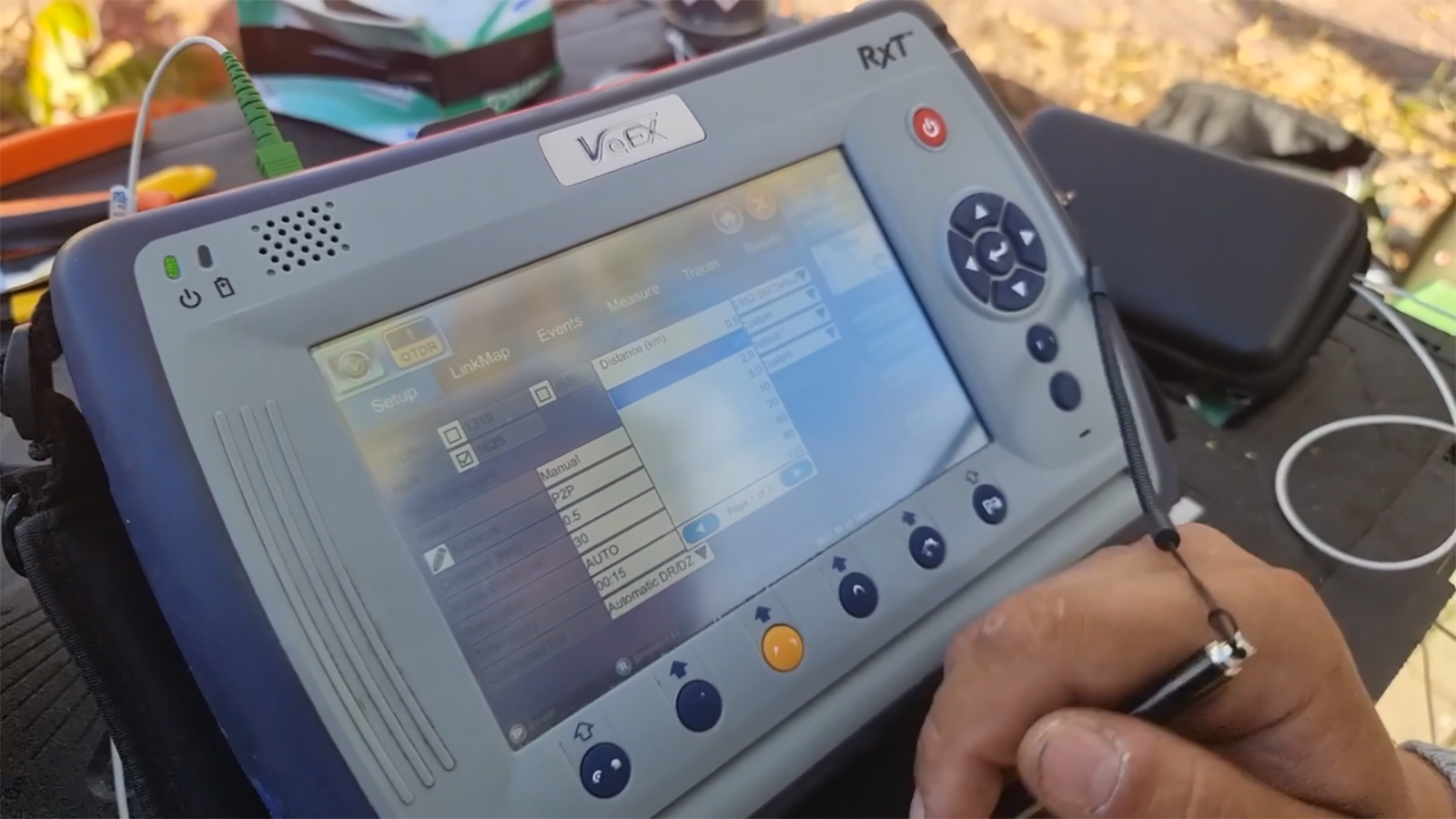 Ilustracja przedstawia urządzenie z dużym ekranem na frontowej ścianie. Ekran jest dotykowy, o czym świadczy rysik, trzymany w dłoni przez osobę obsługującą urządzenie. Pod ekranem oraz obok po prawej znajdują się przyciski. Do górnej ściany przedmiotu przyłączony jest kabel. Nad ekranem znajduje się napis fał e e iks.