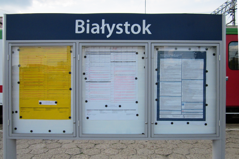 Fotografia przedstawia kolejowe rozkłady jazdy, umieszczone w przeszklonych gablotach, ustawionych na peronie, na dworcu kolejowym.