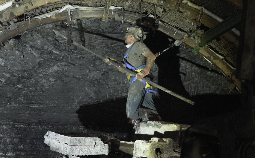 Na zdjęciu widać górnika trzymającego łom górniczy. Górnik jest ubrany w strój roboczy i kask. Górnik znajduje się blisko ociosu. W rękach trzyma łom górniczy. Łom ma długi stalowy trzon. Na końcu trzonu po lewej stronie widać krótkie zaokrąglone zakończenie. Górnik sprawdza łomem opinkę obudowy. 